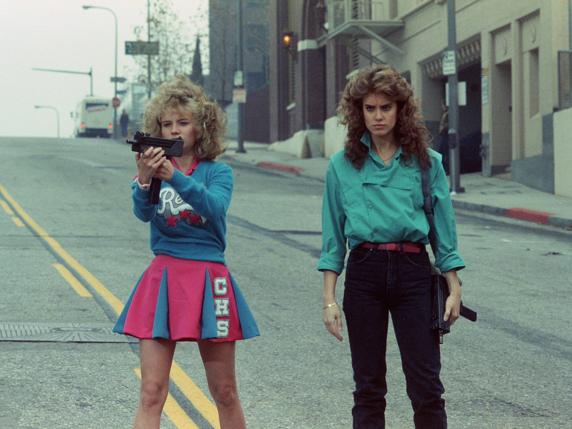 Two women with curly hair standing on empty street. Left wears blue jumper and red cheerleader skirt, right wears teal shirt and dark trousers.