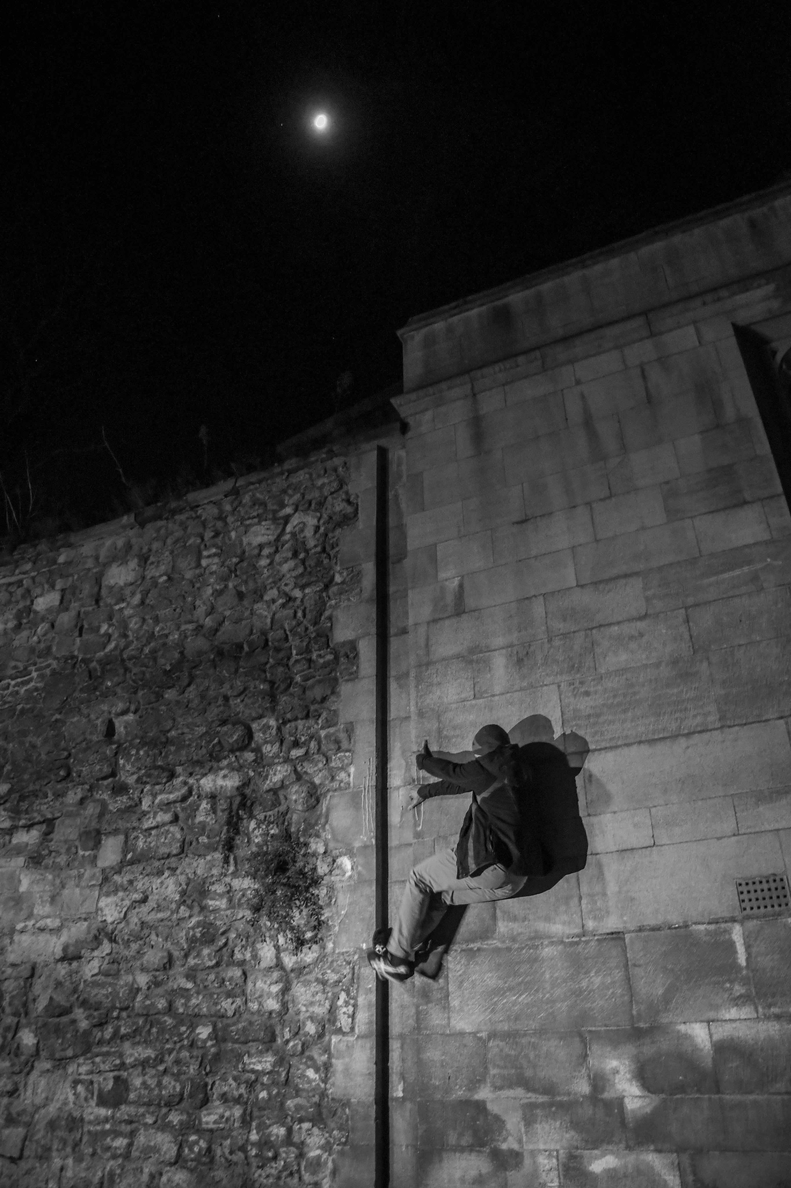 Black and white image showing person climbing pole against brick wall at night, with bright moon visible in dark sky above.