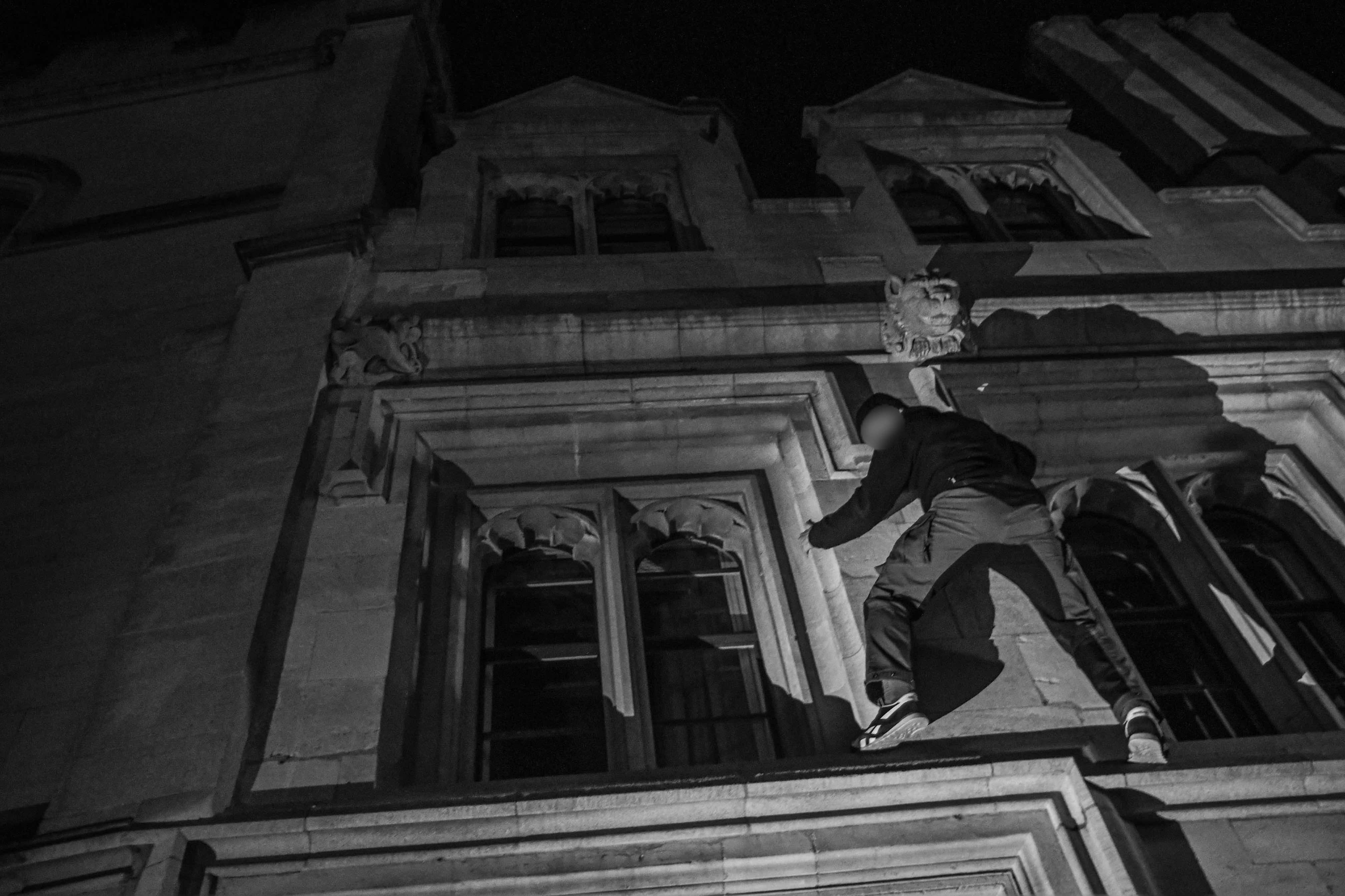 Black and white image of person climbing through ornate building window at night, shot from low angle showing architectural details.