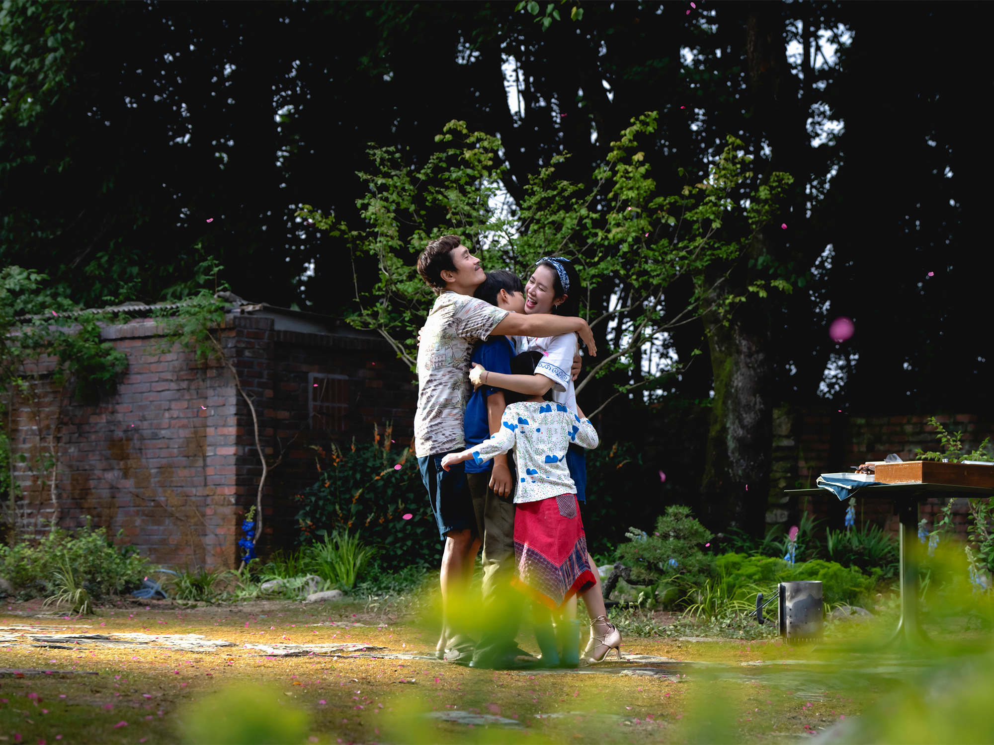 Woman and child embracing in garden with brick wall, green foliage, and wooden table in background.