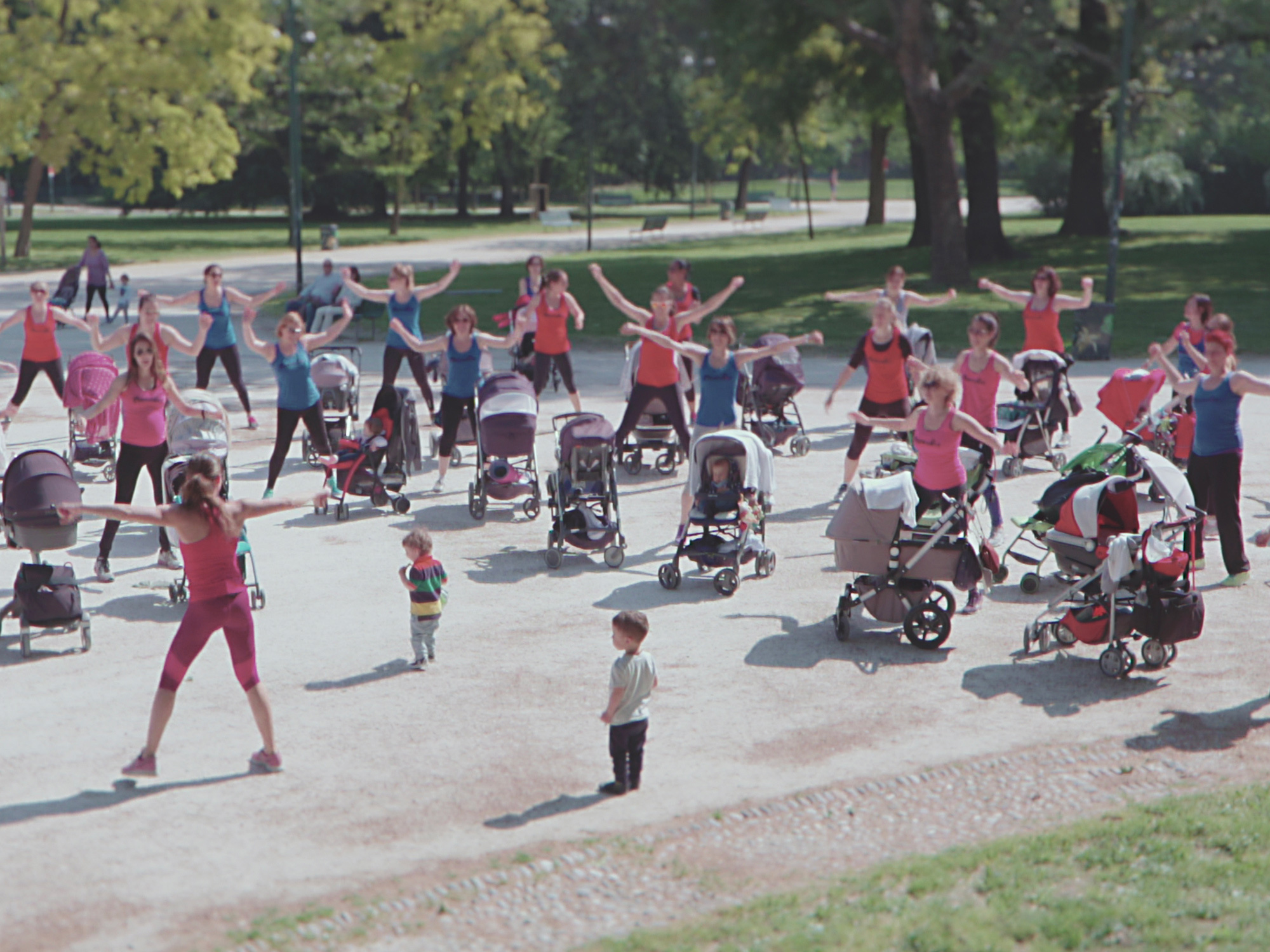 Large group of people in red clothing exercising in a park, with pushchairs and children present.
