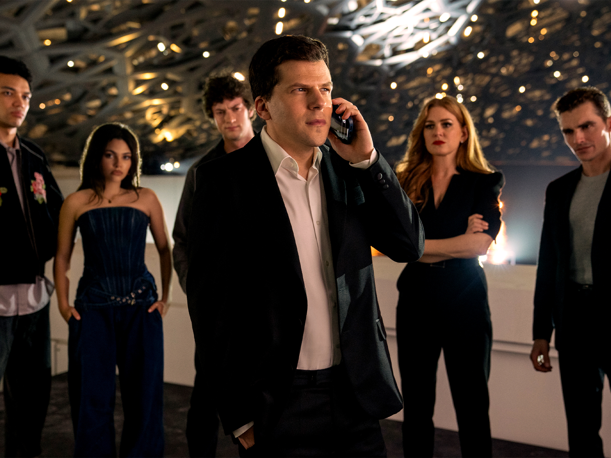Man in dark suit speaking on mobile phone with group of formally dressed people standing behind him in modern interior space.