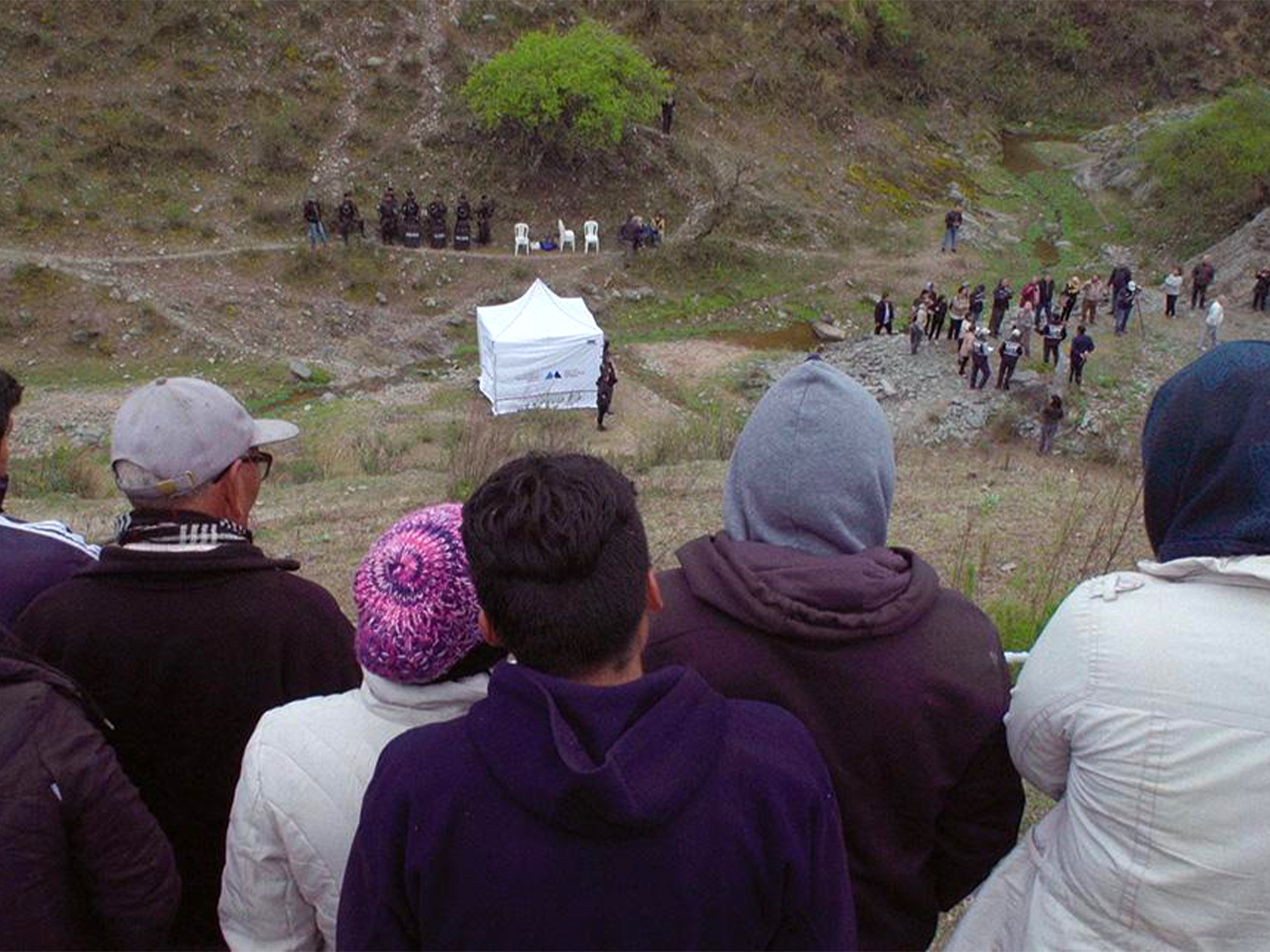 Crowd of people watching from hillside as group gathers around white tent in valley below, surrounded by green vegetation.
