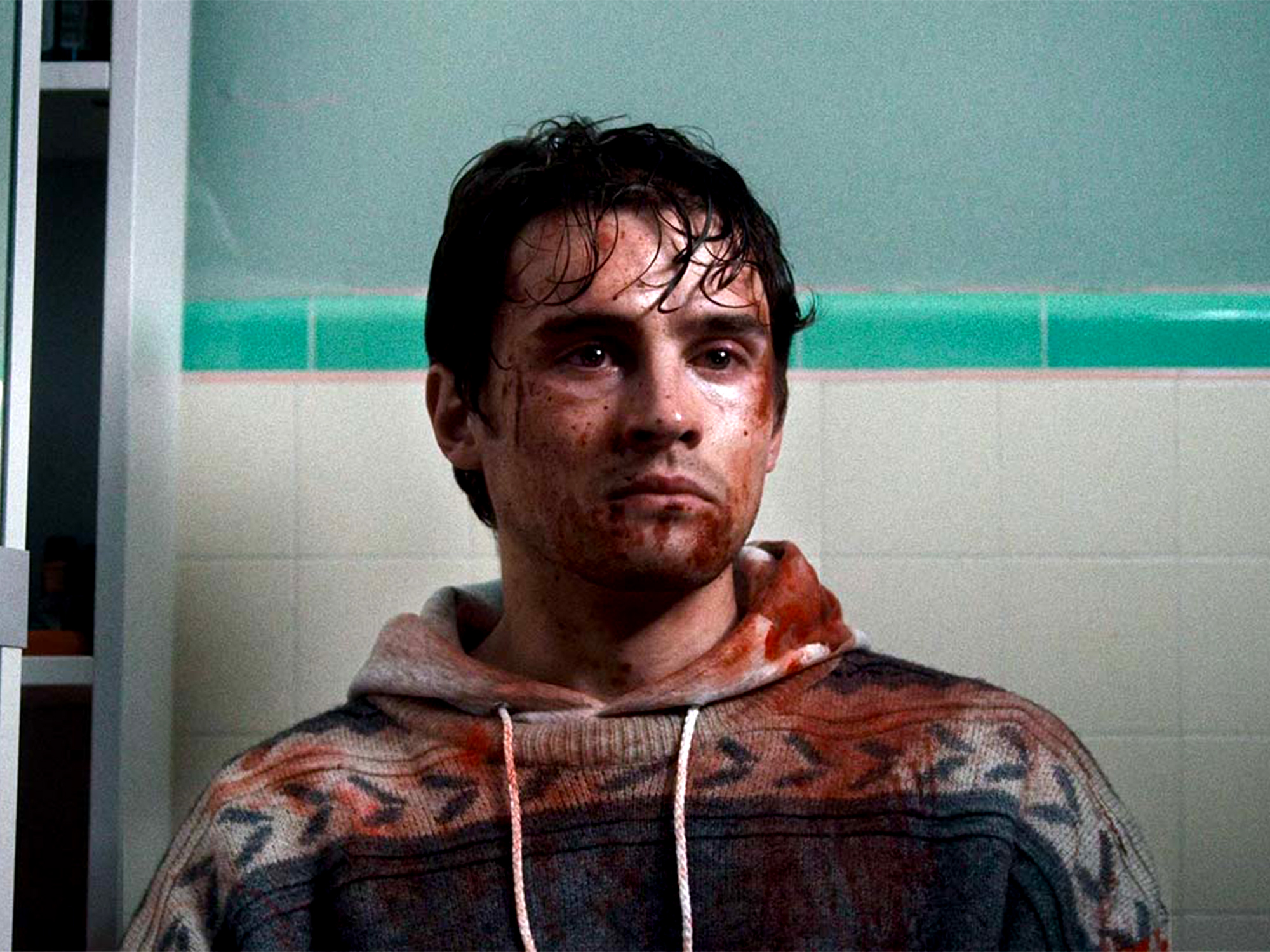 Man with wet dark hair and facial injuries wearing patterned hoodie against mint green and white tiled bathroom wall.