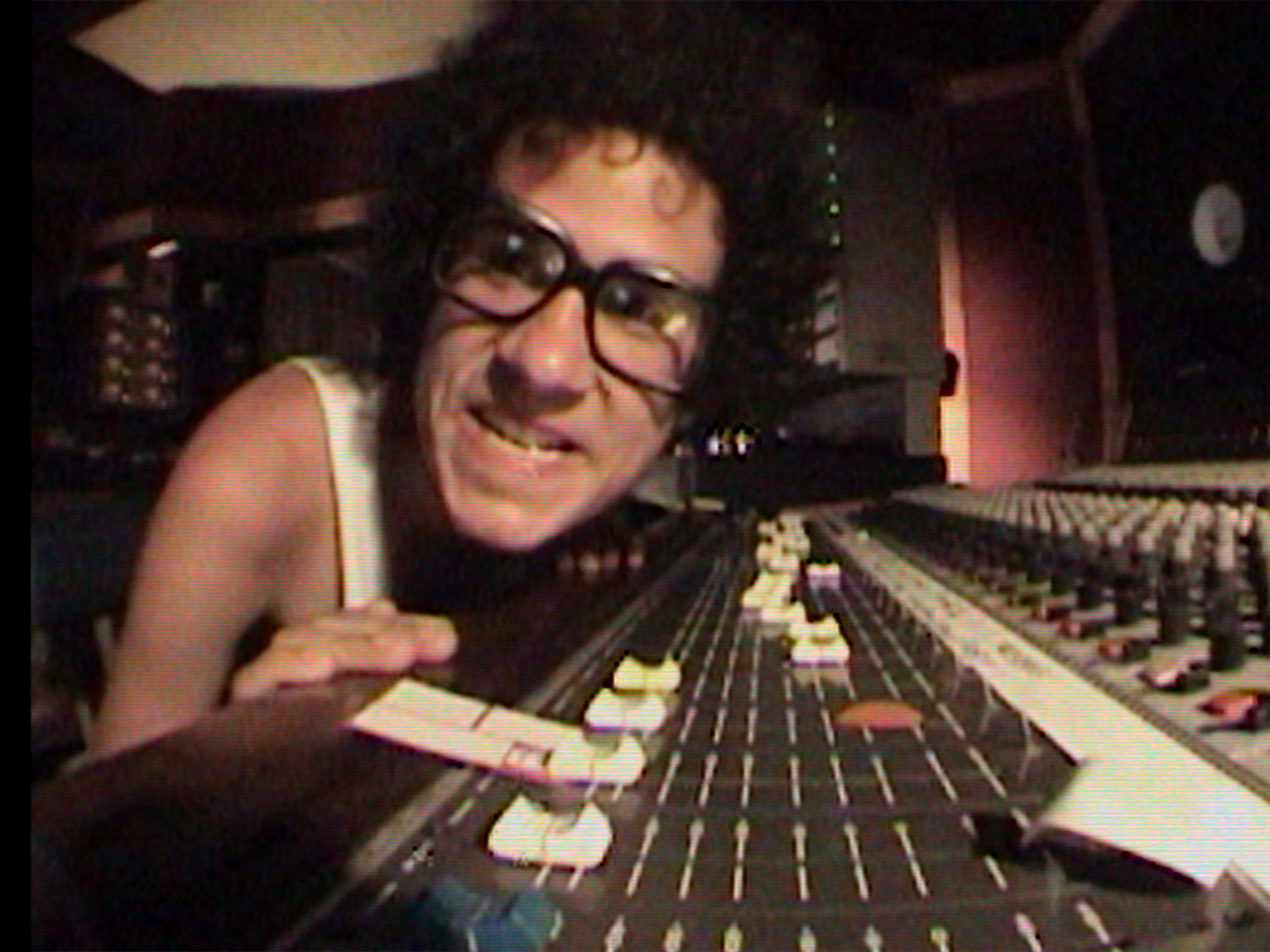Curly-haired person with glasses leaning over large audio mixing console with multiple faders and controls in recording studio.
