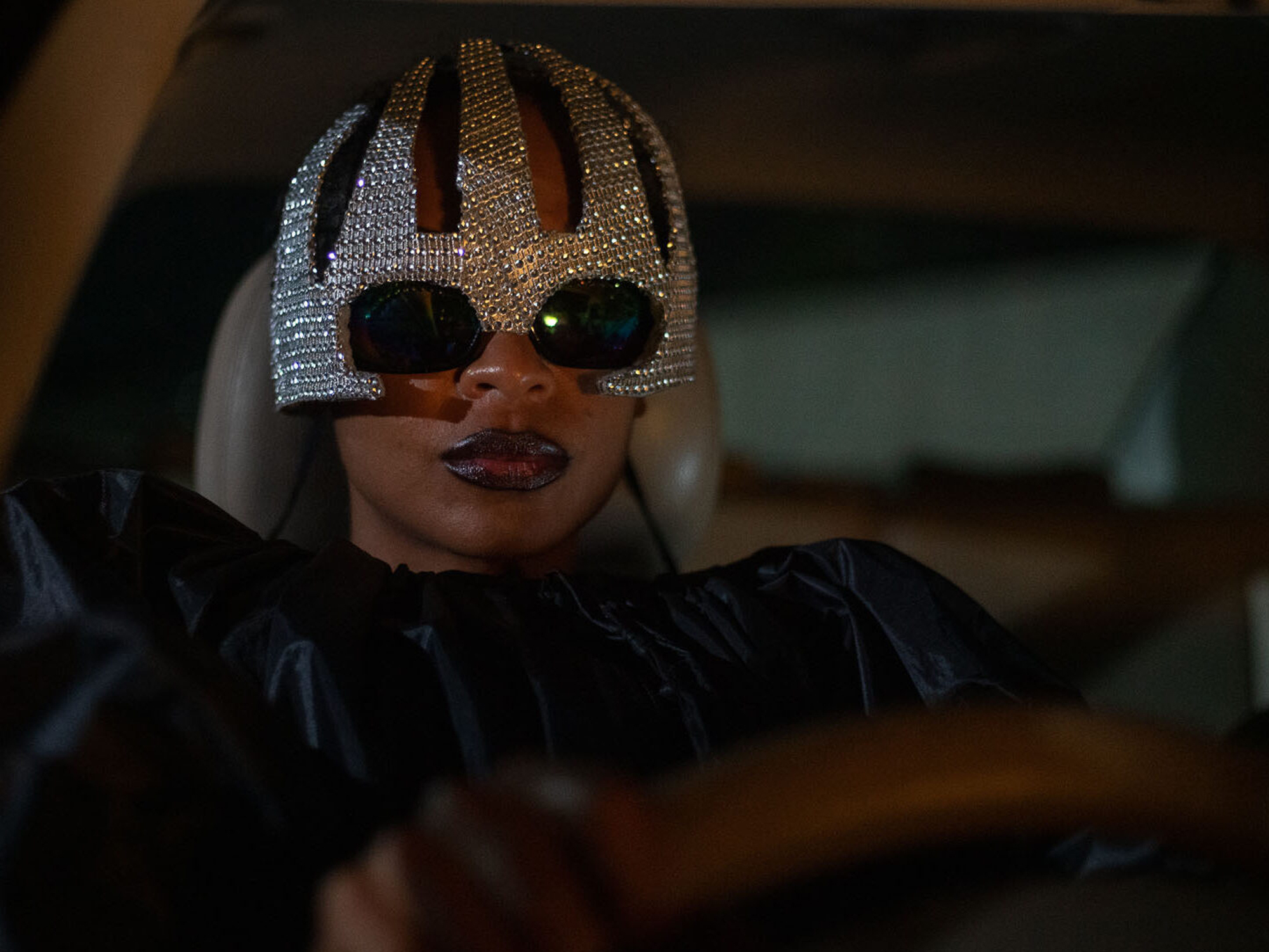 Person wearing elaborate metallic headpiece and sunglasses, in a dark setting