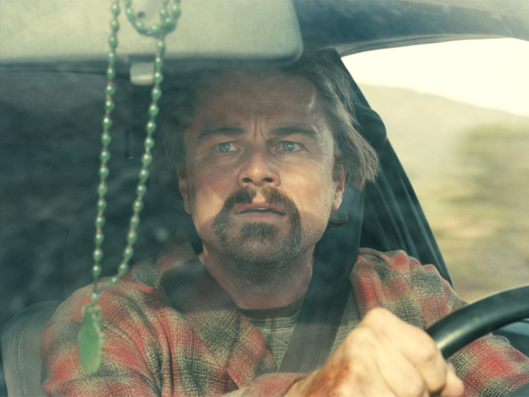 Man with beard driving lorry, wearing red patterned shirt, beaded mirror hanging from roof, view through windscreen.