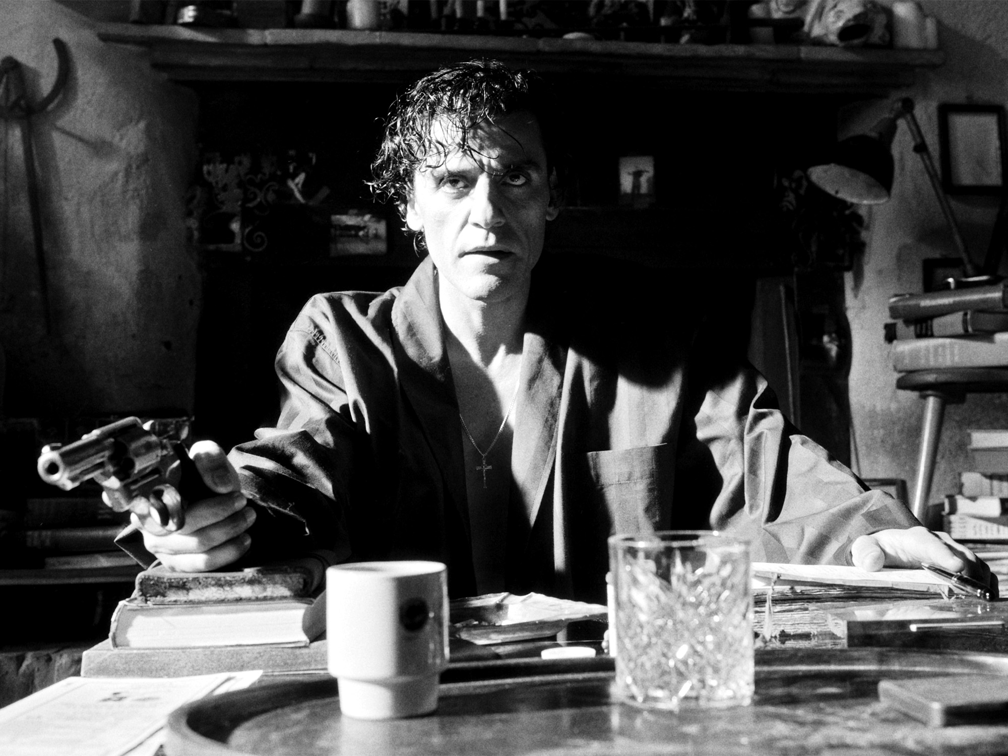 Black and white image of man with curly hair sitting at cluttered table, holding small object, with mug and glass beside scattered papers.