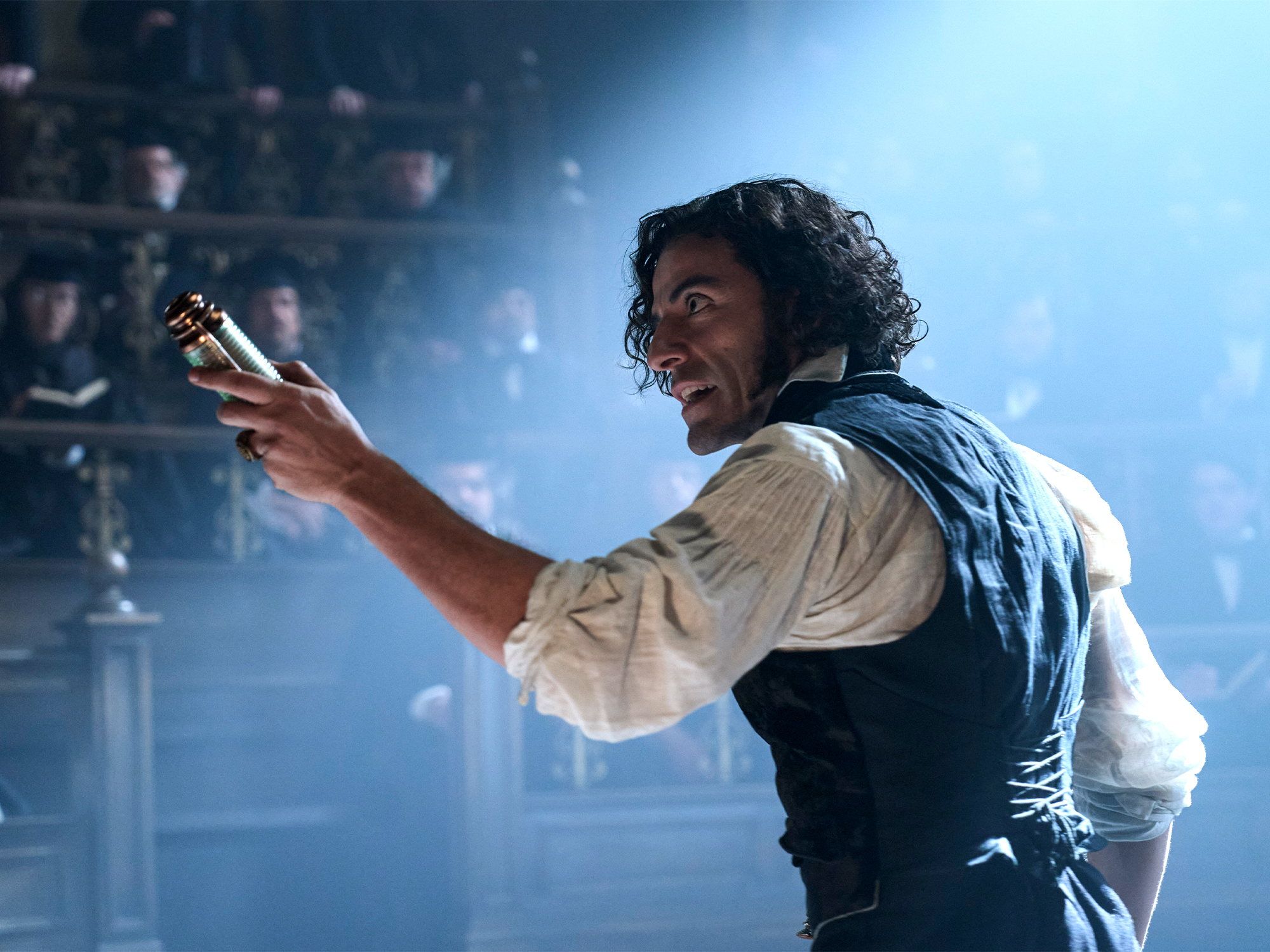 Man with curly hair in white shirt and dark waistcoat holds small object, smiling on stage with blue lighting and audience in background.