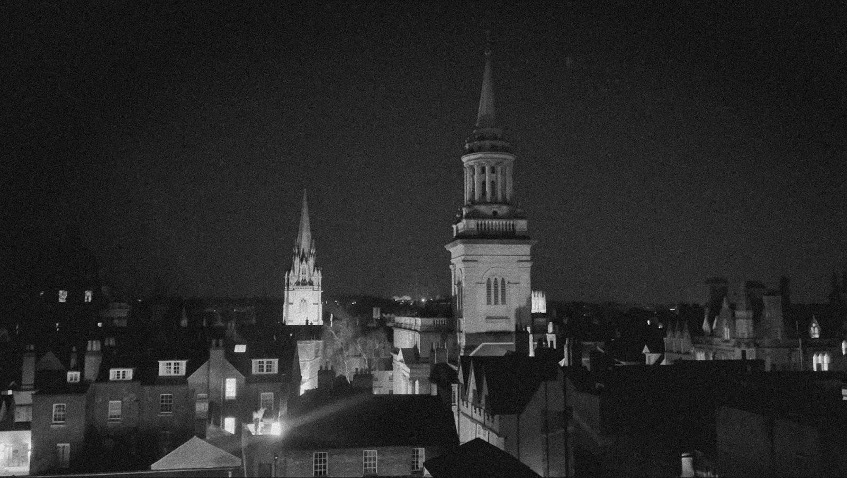 Night view of historic town with illuminated church spire and bell tower rising above residential buildings, black and white.