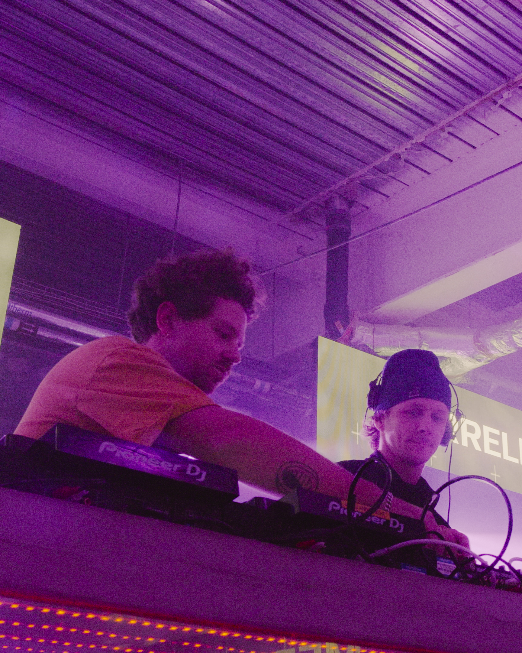 Two men at DJ equipment under wooden ceiling, bathed in purple and pink lighting with visible steam or fog effects.