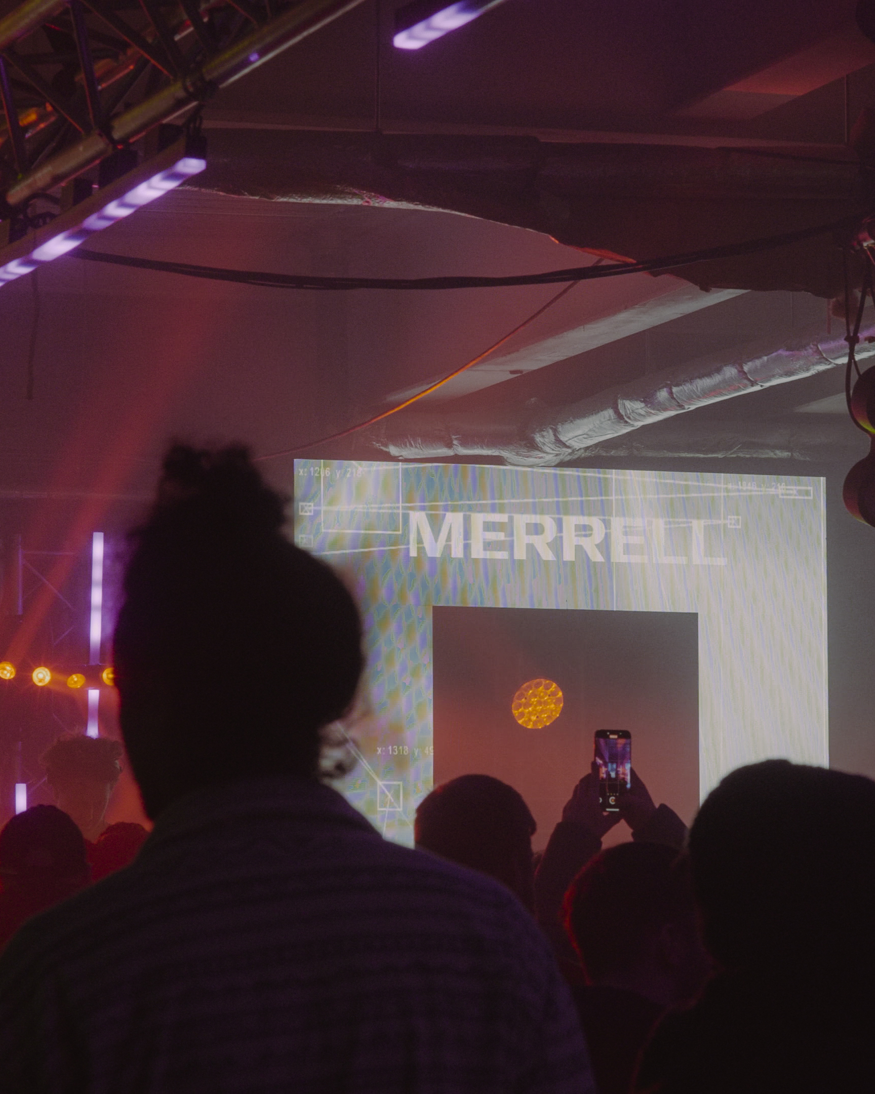 Dark silhouettes of audience members facing illuminated projection screen displaying "MERRELL" text in industrial space with purple lighting.