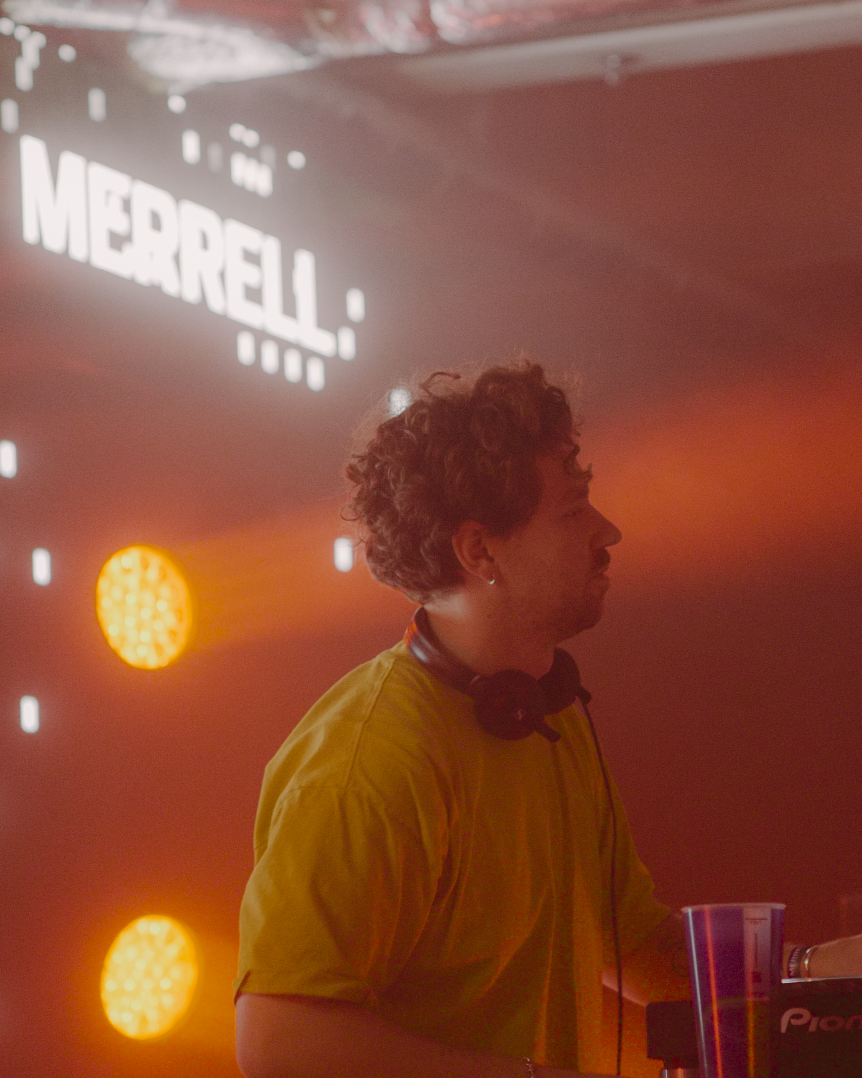 Person with curly hair in yellow shirt at event with red lighting, orange stage lights, and "MIXMAG" projection visible in background.