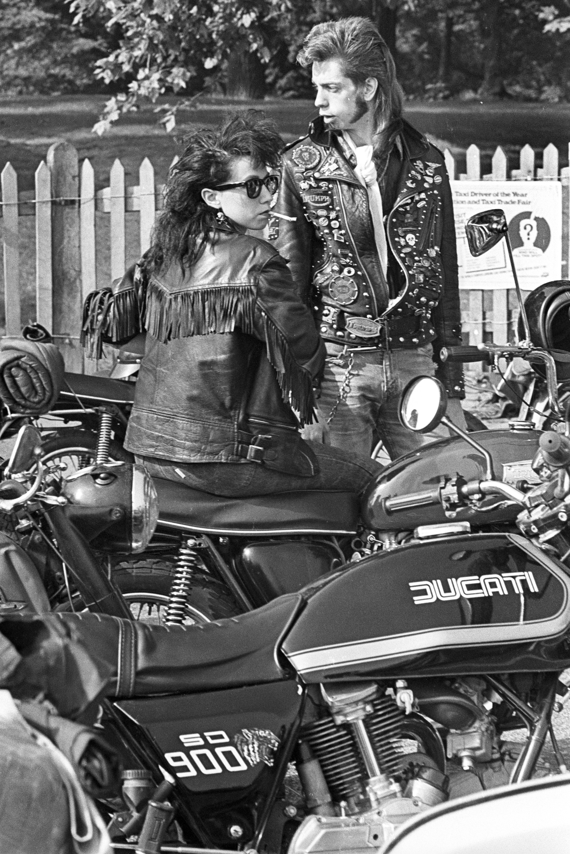 Black and white image of two people in leather jackets beside a Ducati 900 motorcycle, with white picket fence and trees in background.