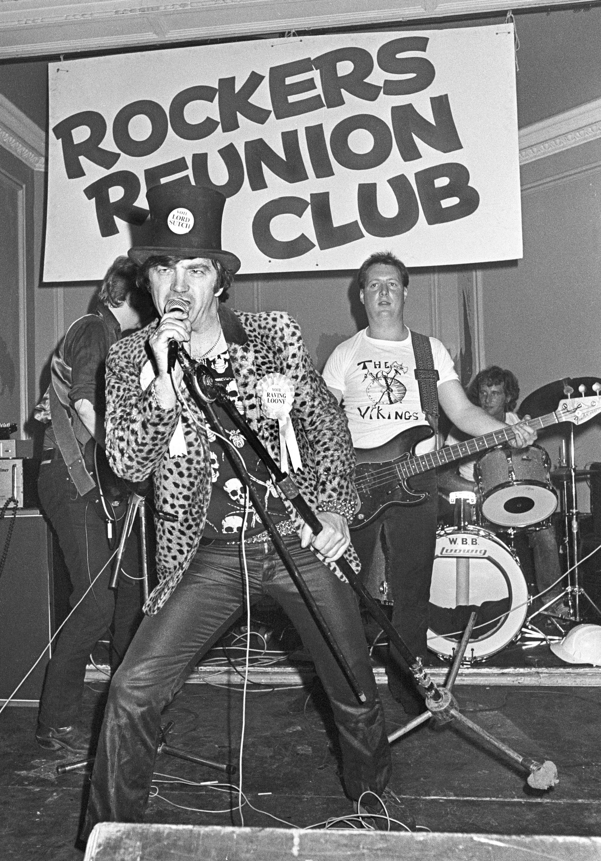 Black and white image of singer in leopard print jacket and top hat performing with microphone beneath "Rockers Reunion Club" sign, band members visible behind.