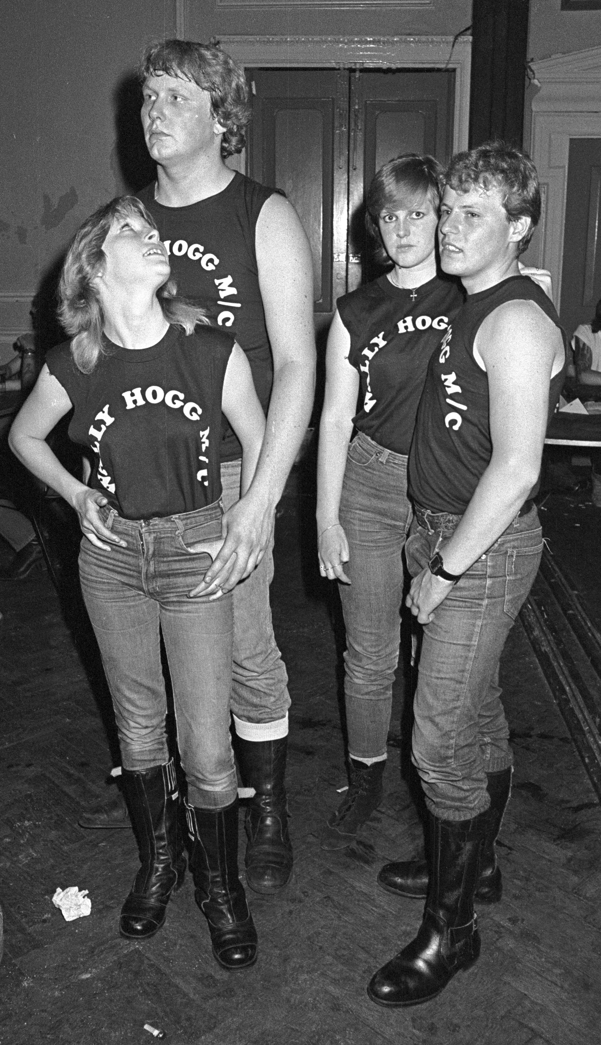 Four people in matching black vests with white text, wearing jeans and boots, standing together indoors. Black and white photograph.