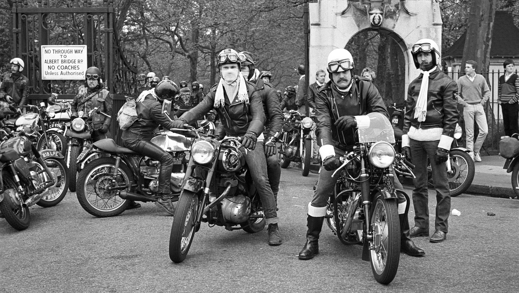 Black and white image of motorcyclists gathered near stone archway, wearing helmets and leather gear, with spectators and bare trees in background.