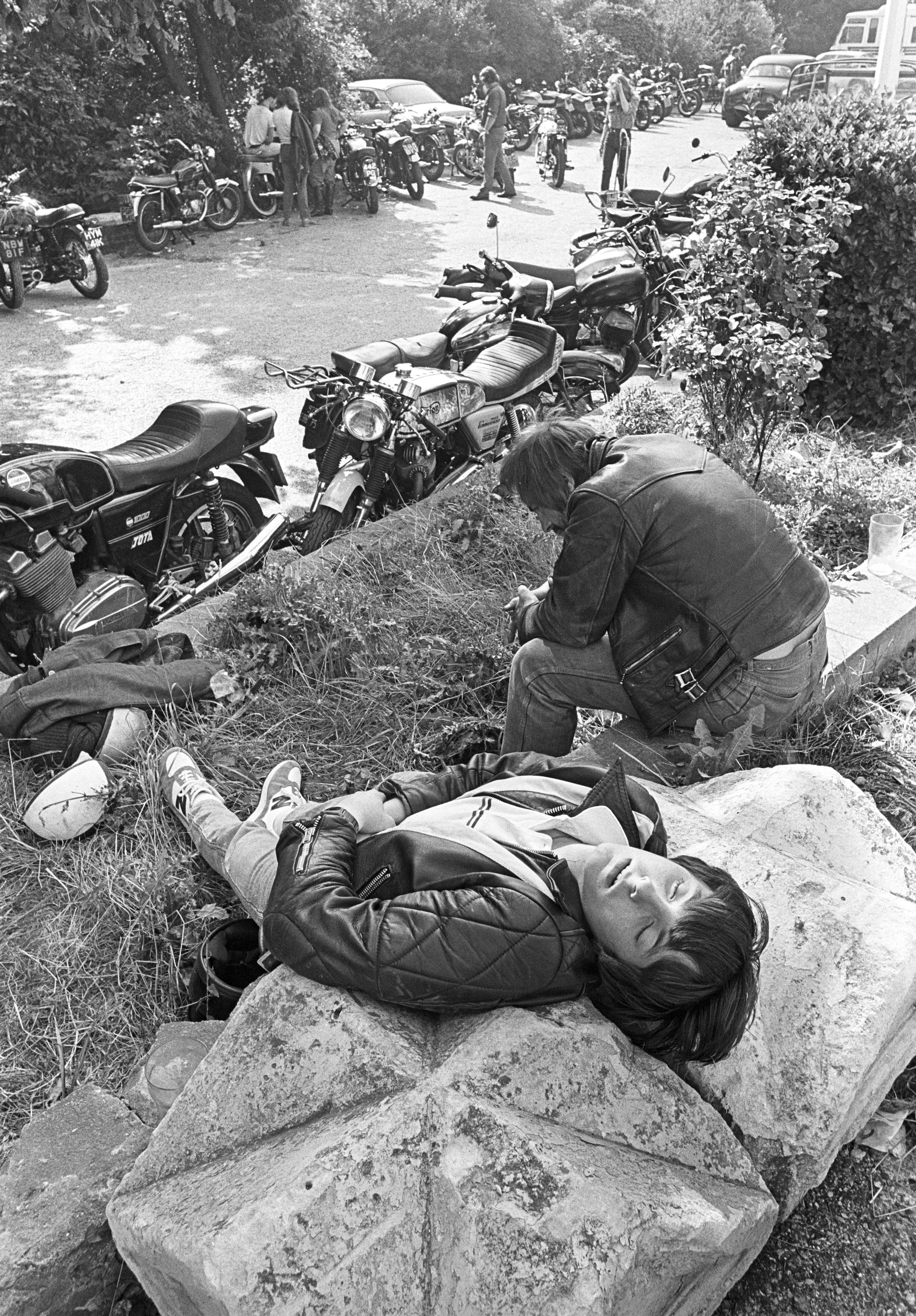 Black and white image showing person lying on stone steps whilst another kneels beside them, with motorcycles and people gathered on road above.