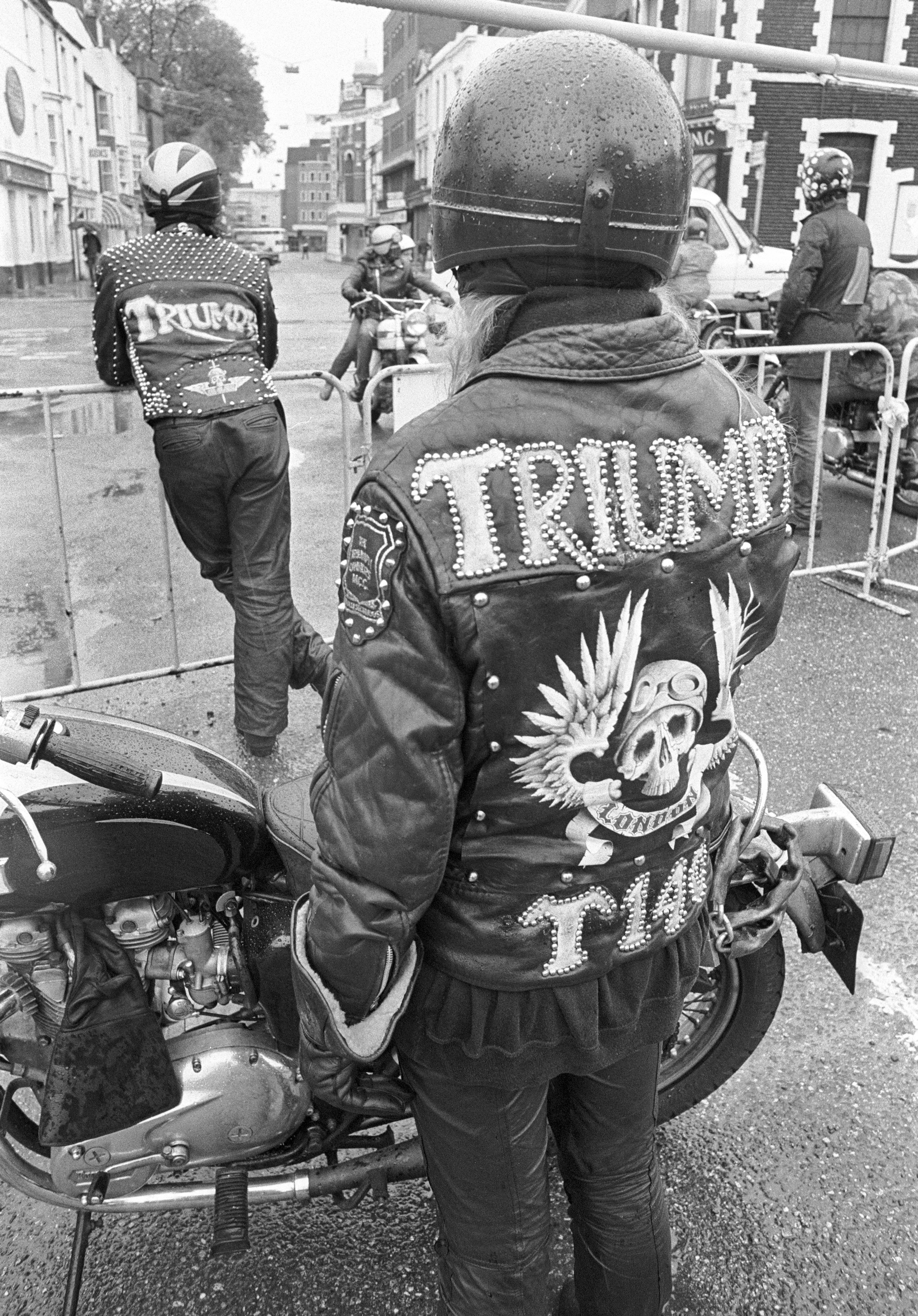 Black and white image of motorcyclist wearing studded leather jacket with "TRIUMPH" text and winged skull design, seated on bike in street scene.