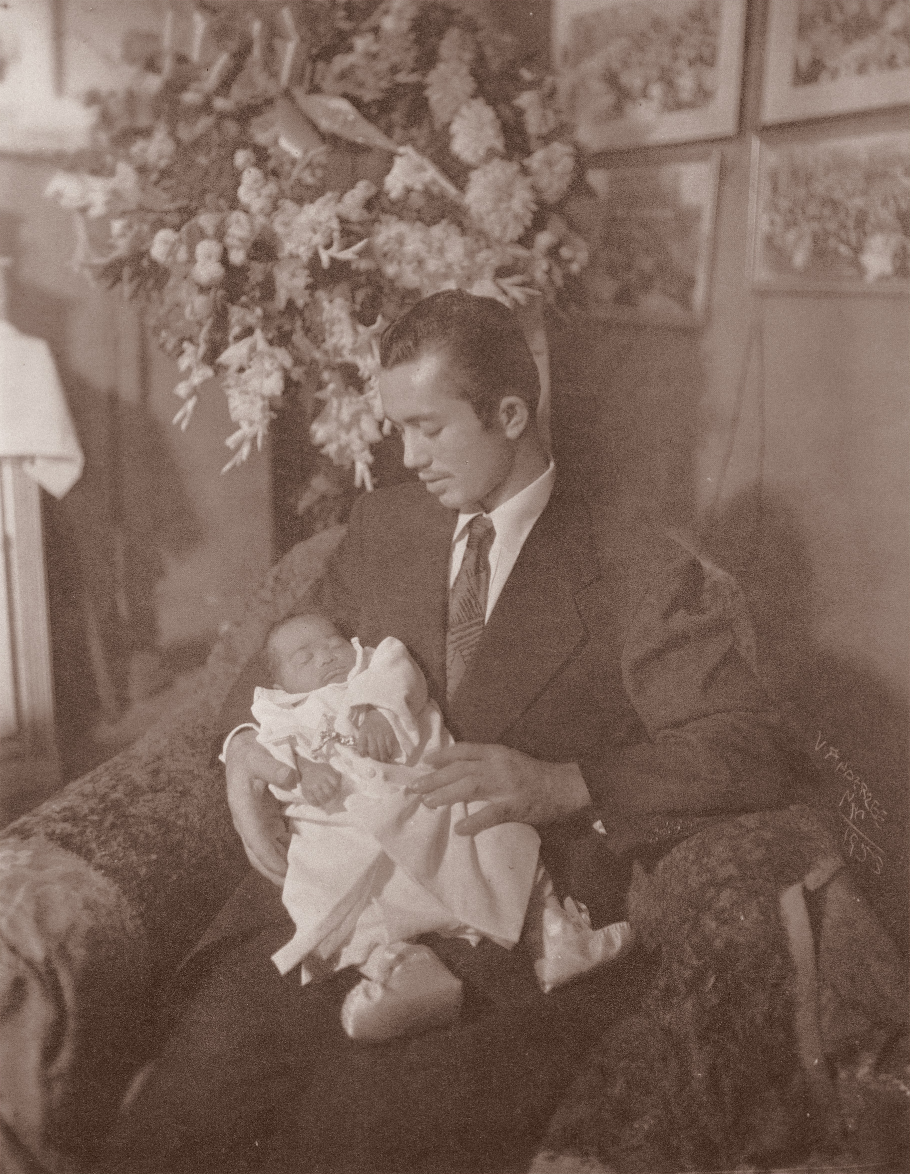 Man in suit and tie holding infant in white christening gown, seated indoors with large floral arrangement and framed pictures behind.