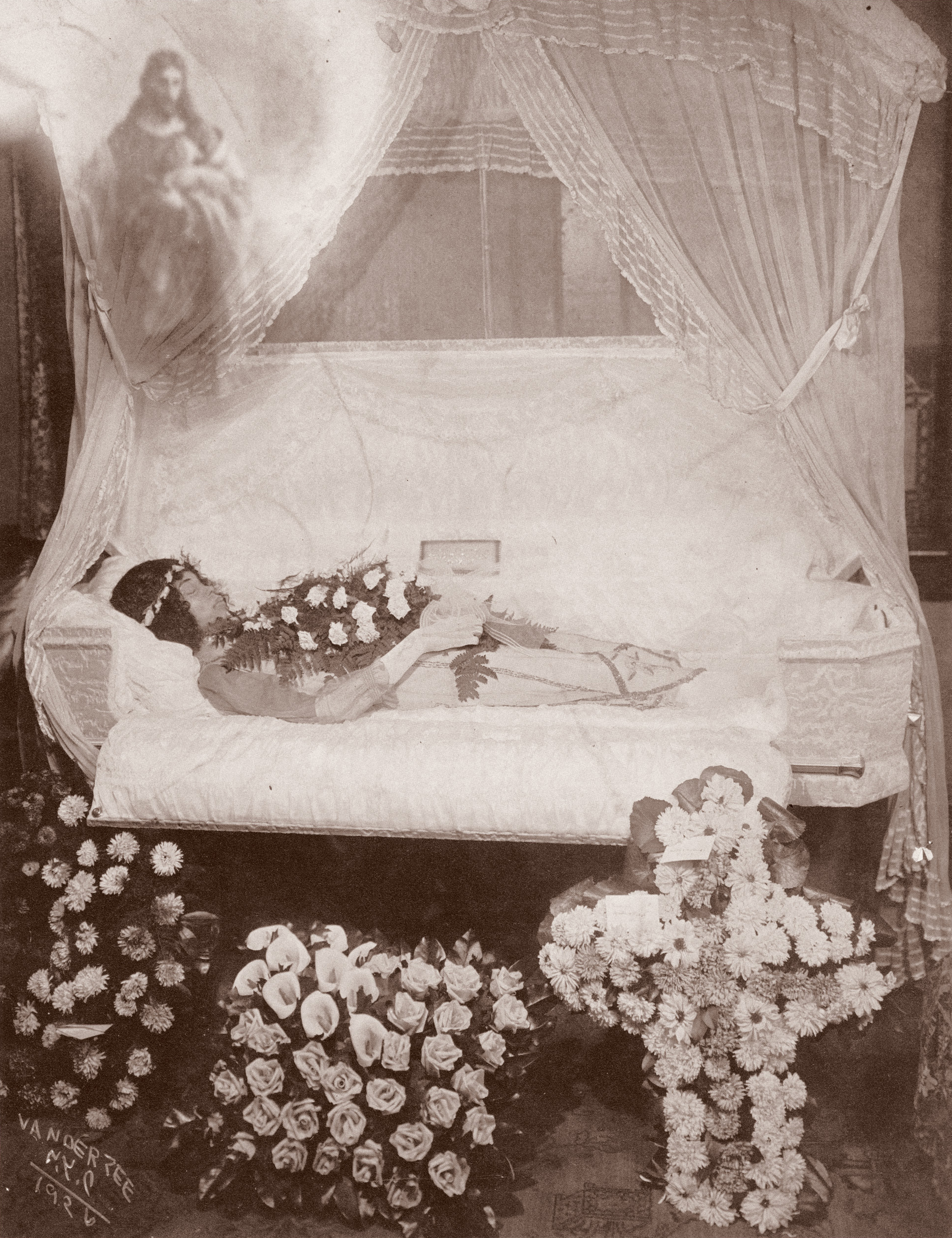 Vintage sepia photograph showing person lying in ornate bed with canopy curtains, surrounded by numerous floral arrangements below.