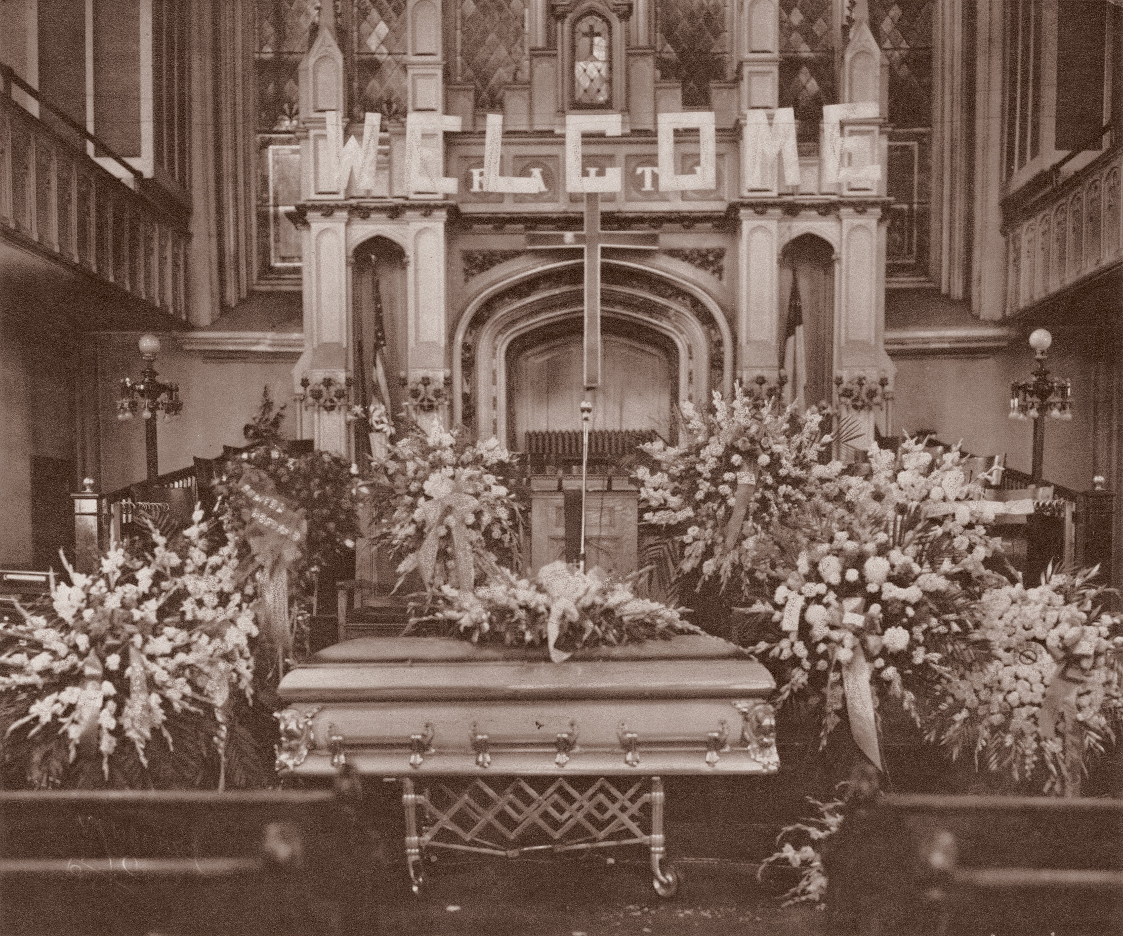 Gothic church interior with ornate casket surrounded by large floral arrangements, arched ceiling, columns, and wooden pews visible.
