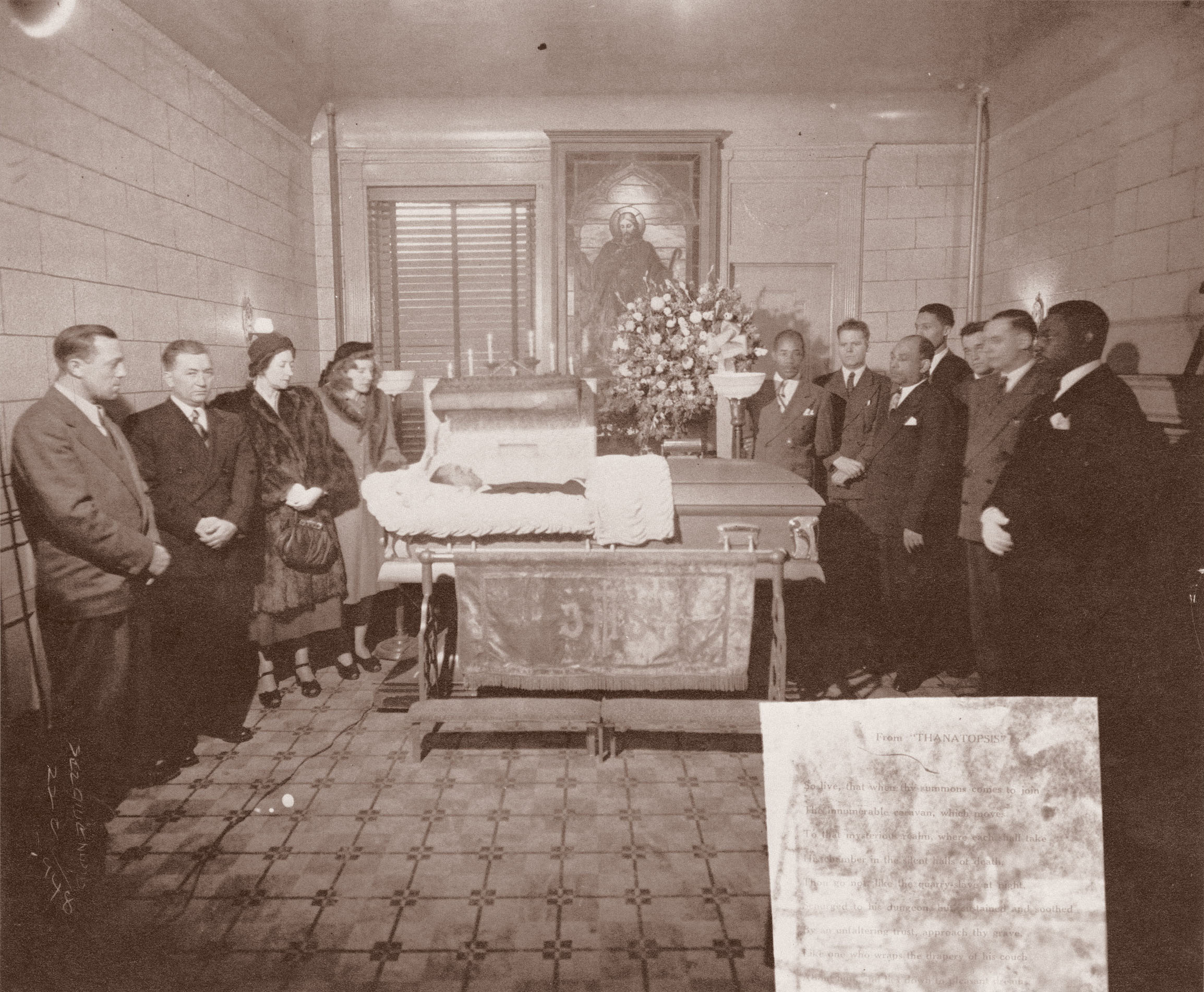 Black and white image showing formally dressed people gathered around a table with flowers in an ornate room with patterned floor.