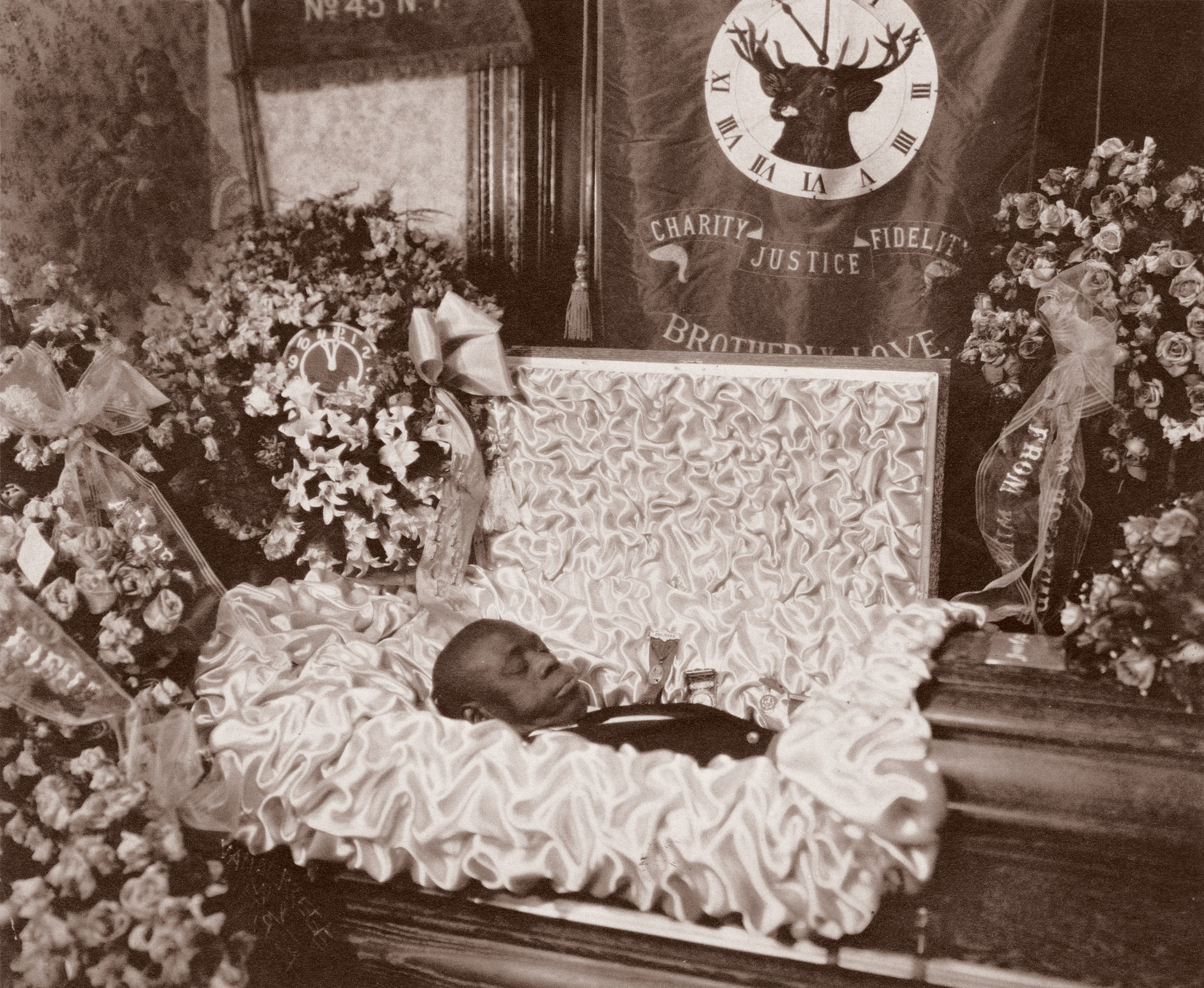 Sepia-toned image of open coffin with deceased person surrounded by floral arrangements, clock on wall showing deer silhouette.