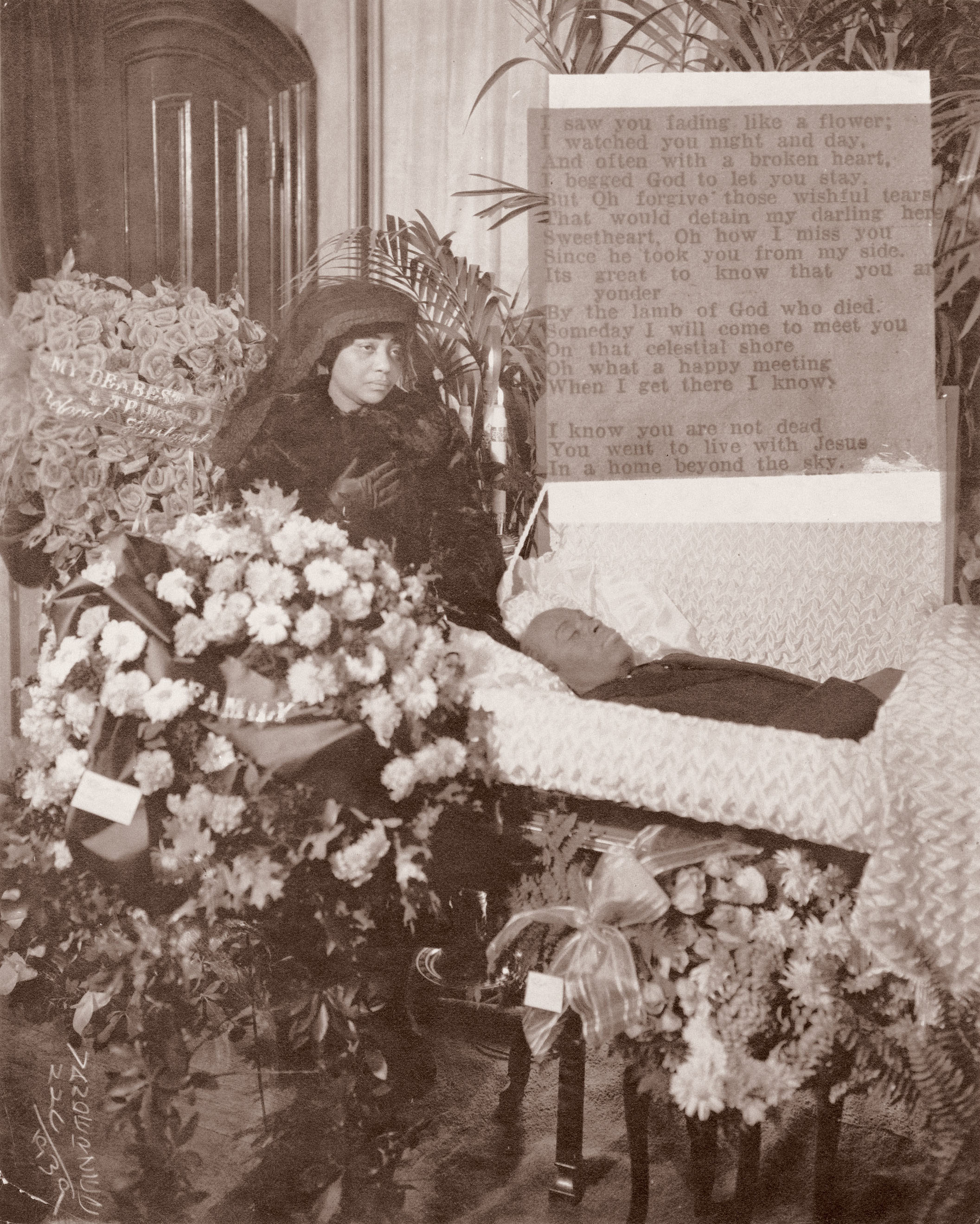 Sepia-toned image of veiled woman in dark clothing kneeling beside memorial plaque, surrounded by large floral arrangements and potted plants.