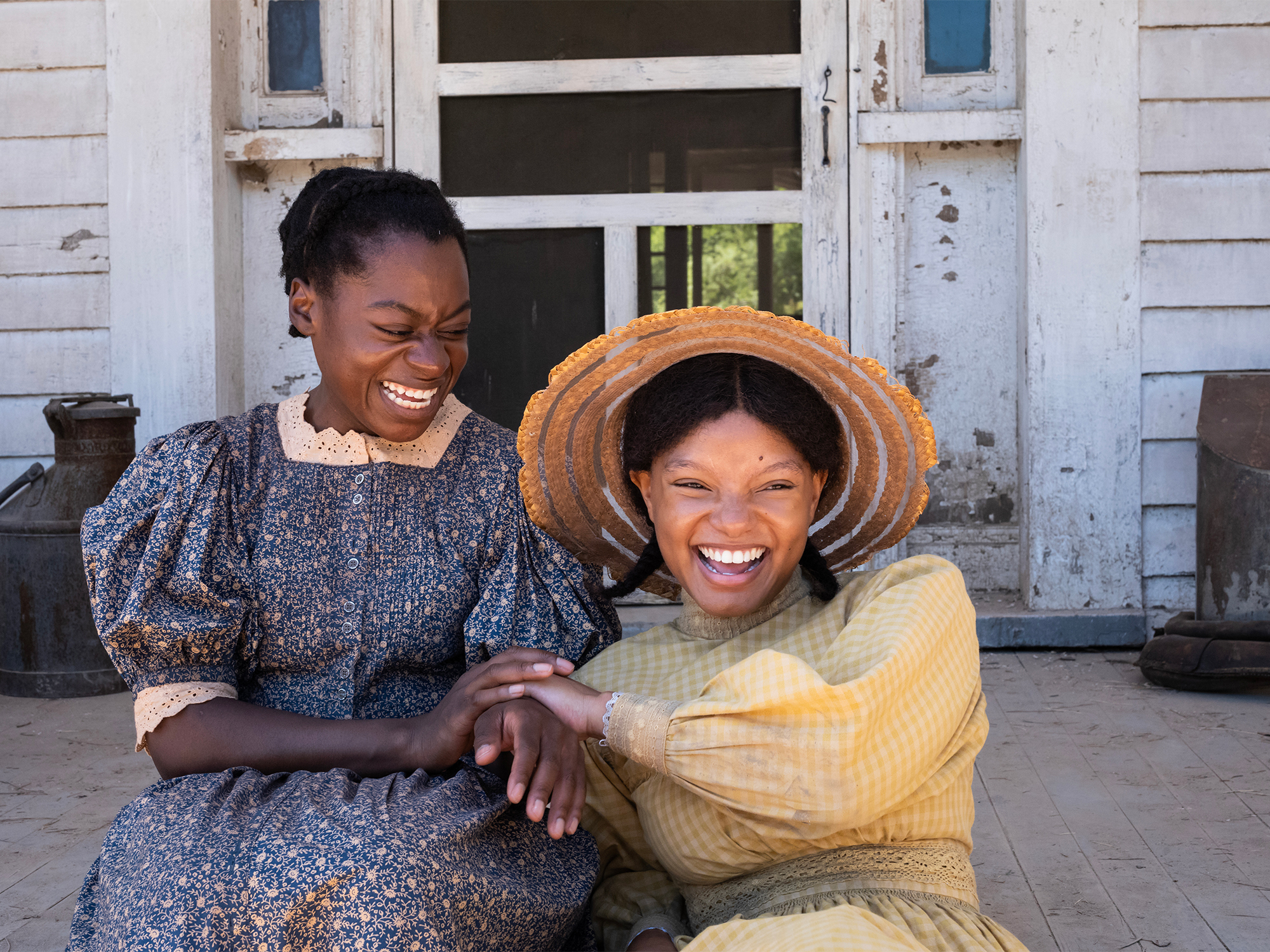 Two smiling women, one wearing a patterned dress, the other a large straw hat, sitting together and laughing.