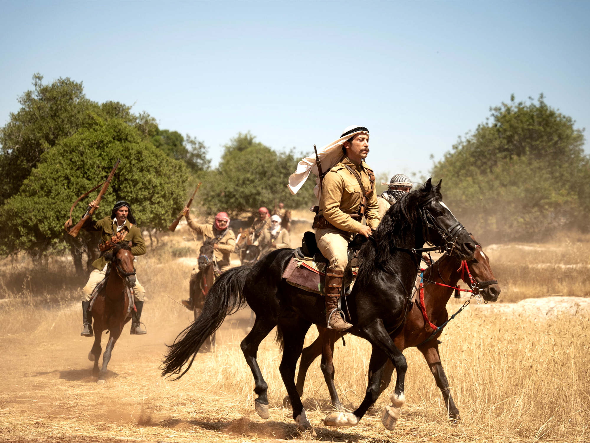 Riders on horseback in desert landscape with trees, dust clouds, wearing traditional Middle Eastern clothing and headwear.
