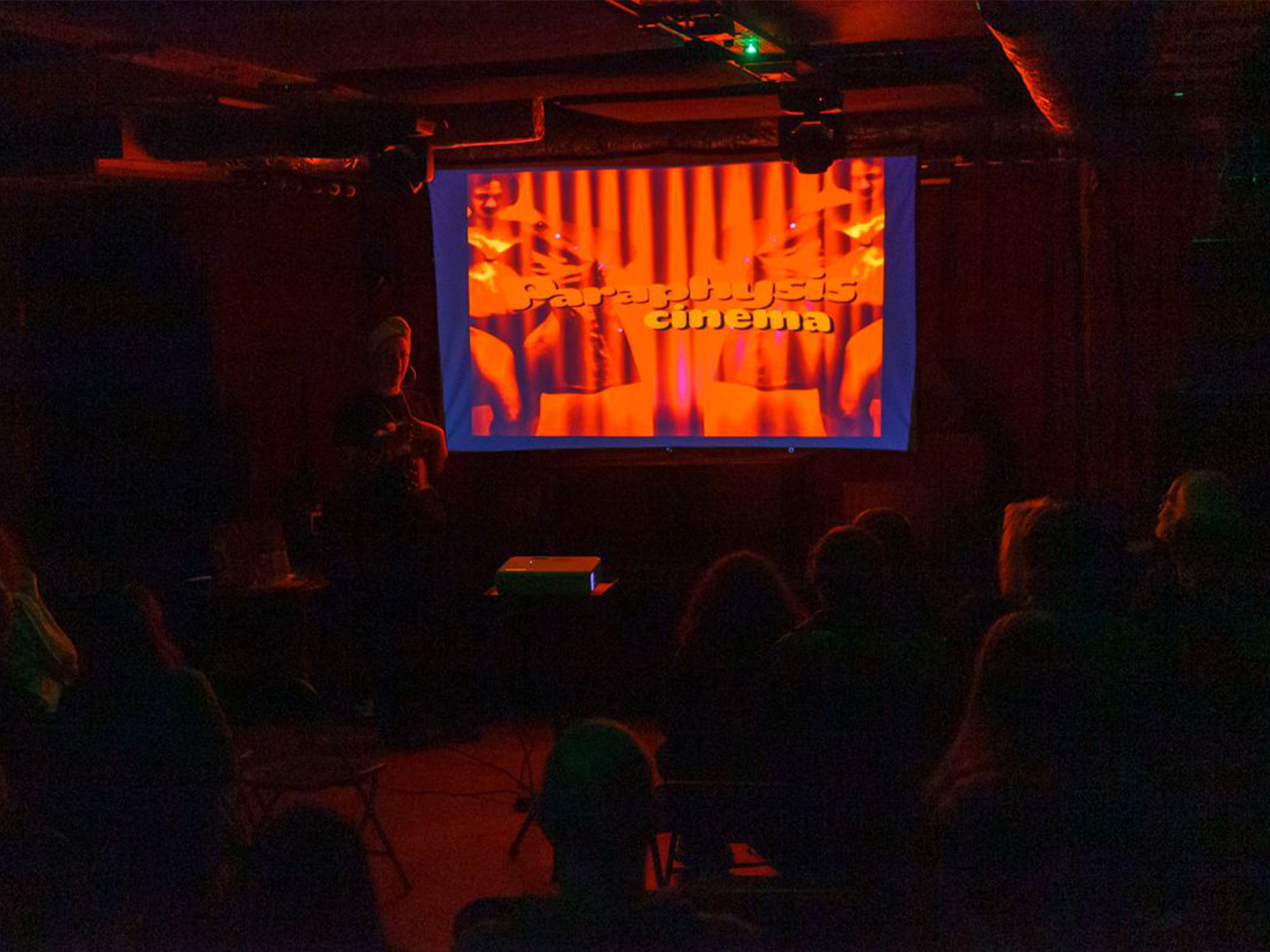 Cinema screen displaying orange and red film title graphics with silhouetted audience members seated in darkened theatre with red lighting.