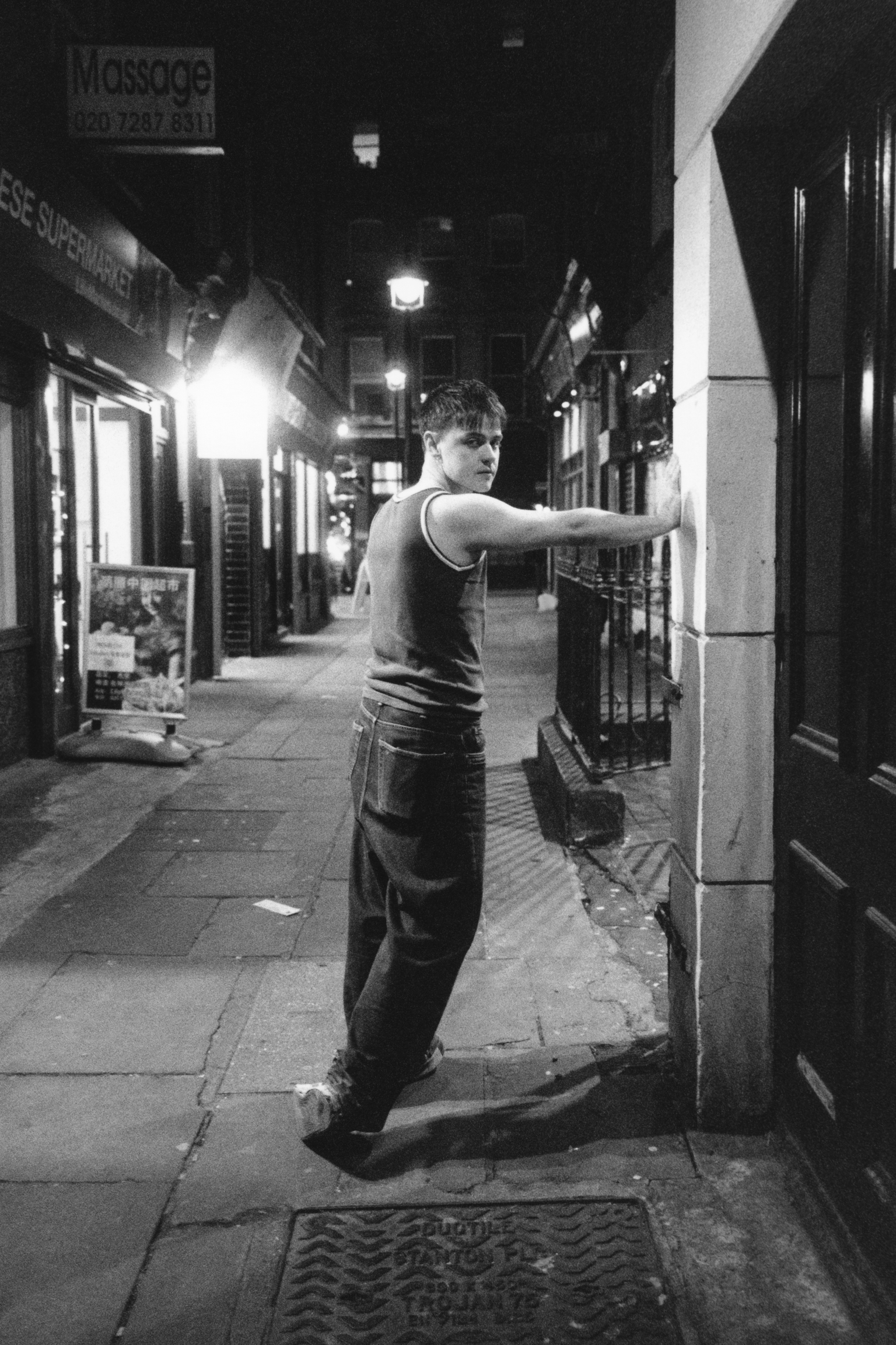 Man in vest stretching against white pillar on night-time street with shops, massage parlour sign visible in background.