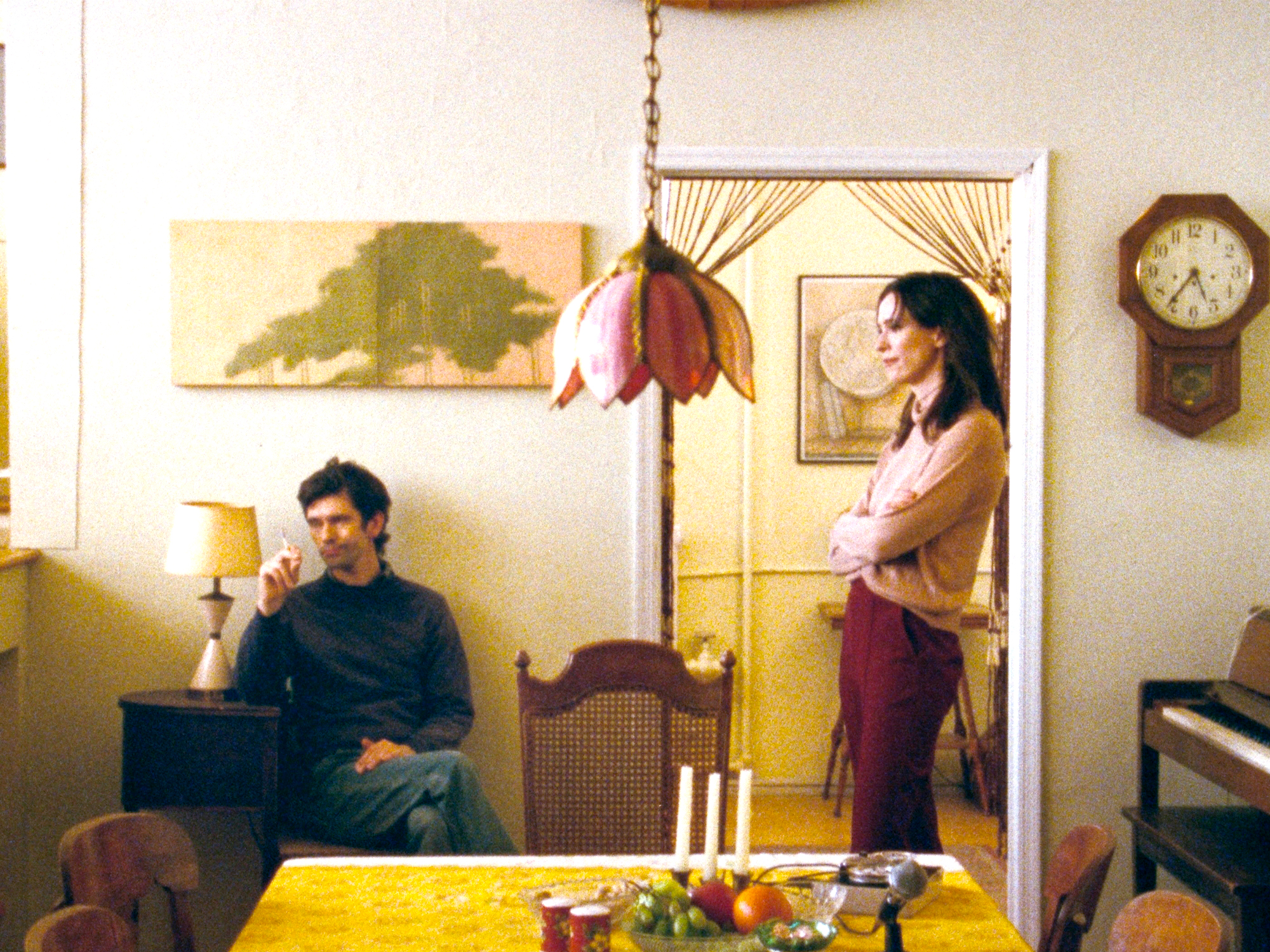 Man in dark jumper sits in armchair whilst woman in brown cardigan stands by doorway in warm-lit living room with yellow tablecloth.
