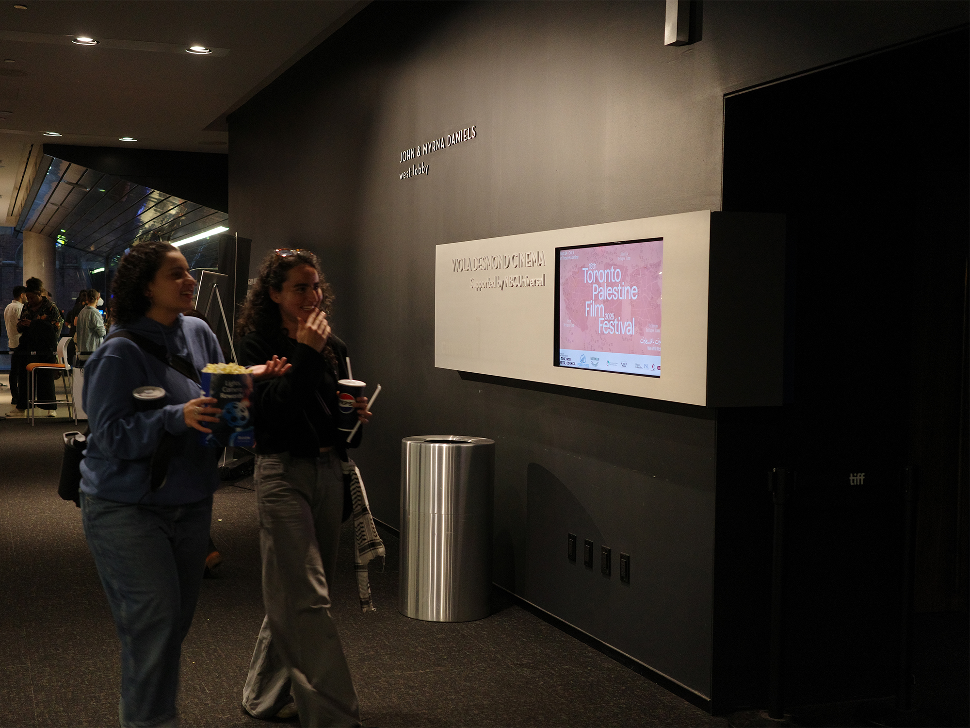 Two people standing in modern gallery space with grey walls, viewing mounted screen displaying pink content next to cylindrical metal bin.