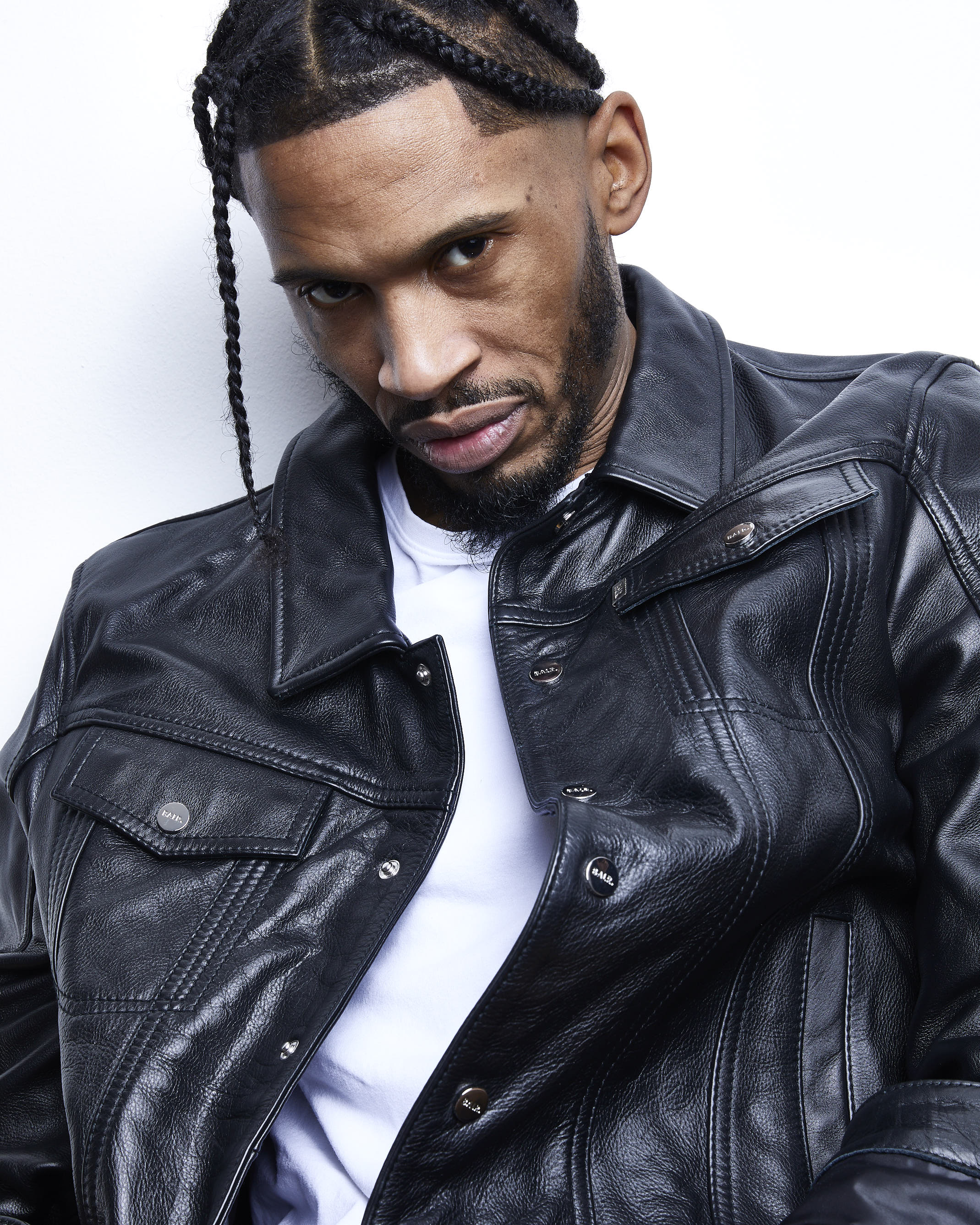 Man with braided hair wearing black leather jacket over white t-shirt, looking directly at camera against white background.