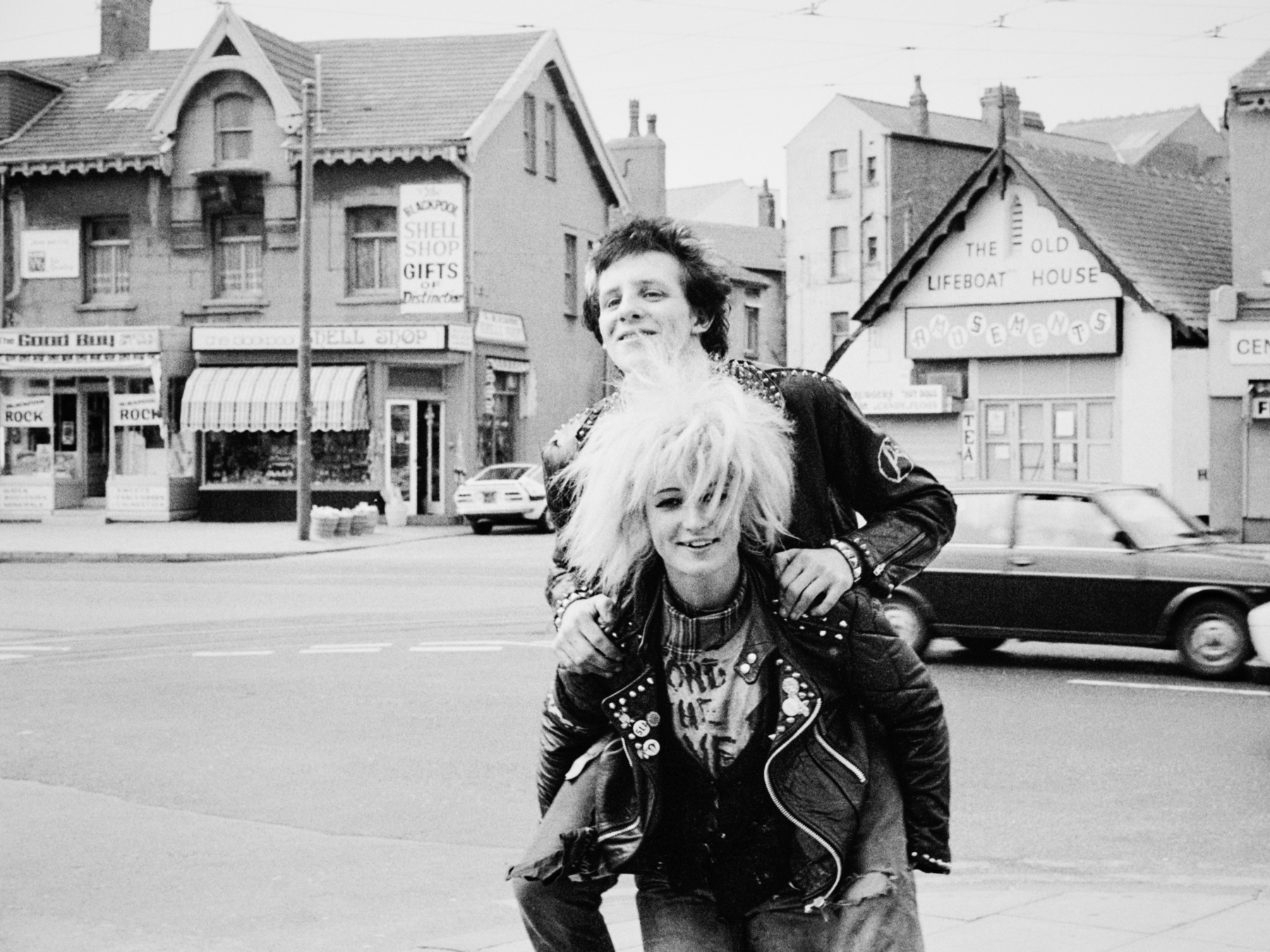 Two people in leather jackets on street, one carrying the other piggyback. Victorian terraced houses and vintage cars in background.