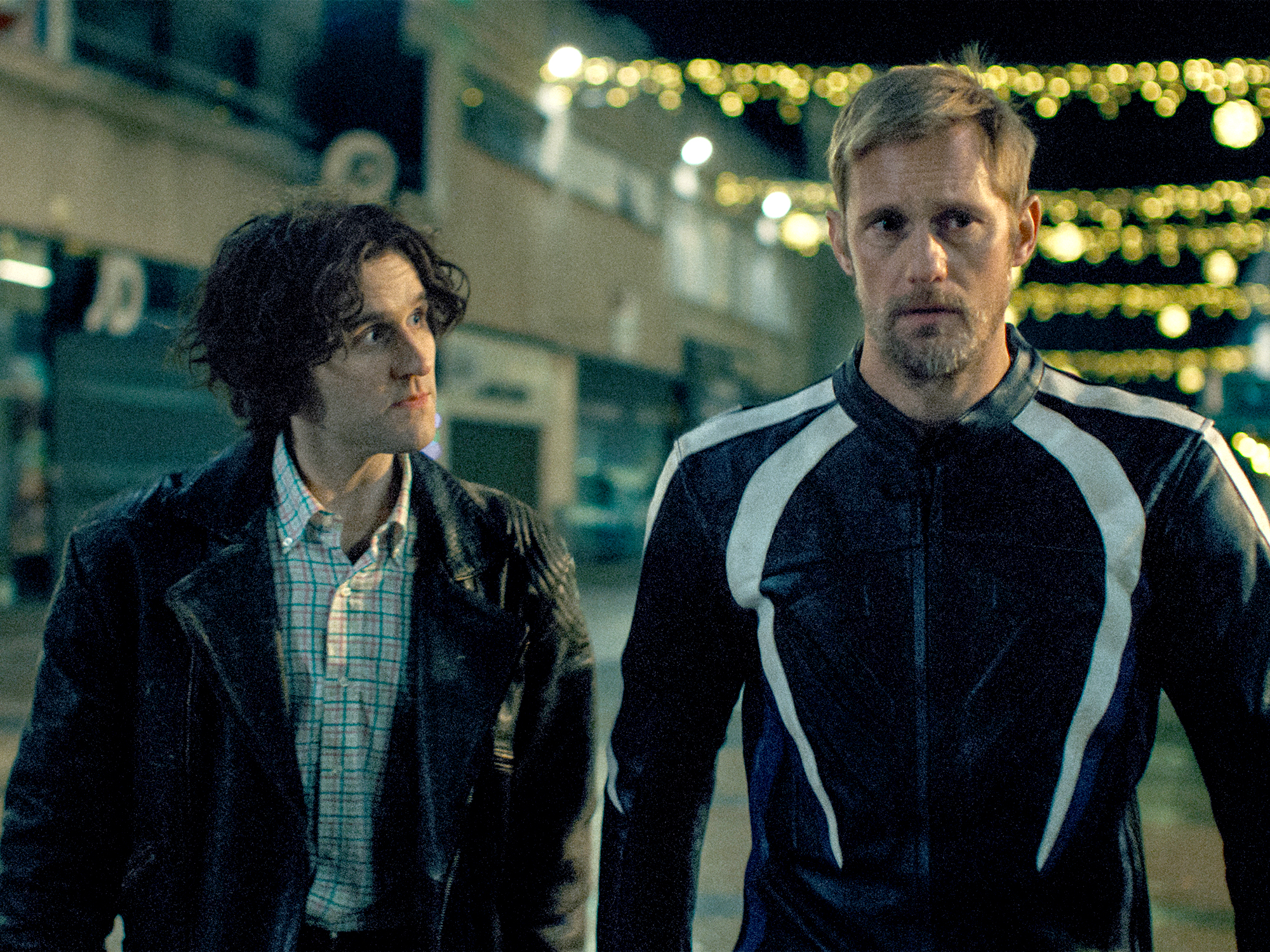 Two men standing outdoors at night - one in black leather jacket over checked shirt, other in black and white athletic top with string lights behind them.