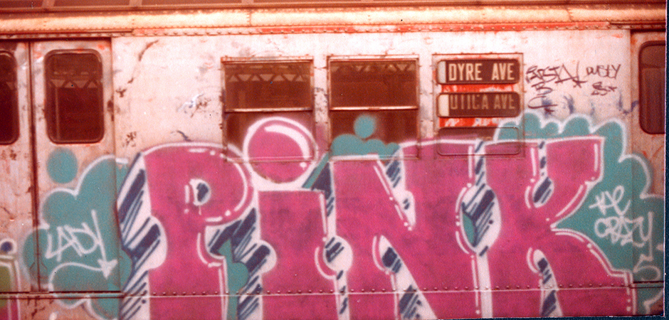 Train carriage covered in large pink graffiti letters with teal and white accents, windows and signage visible above the artwork.