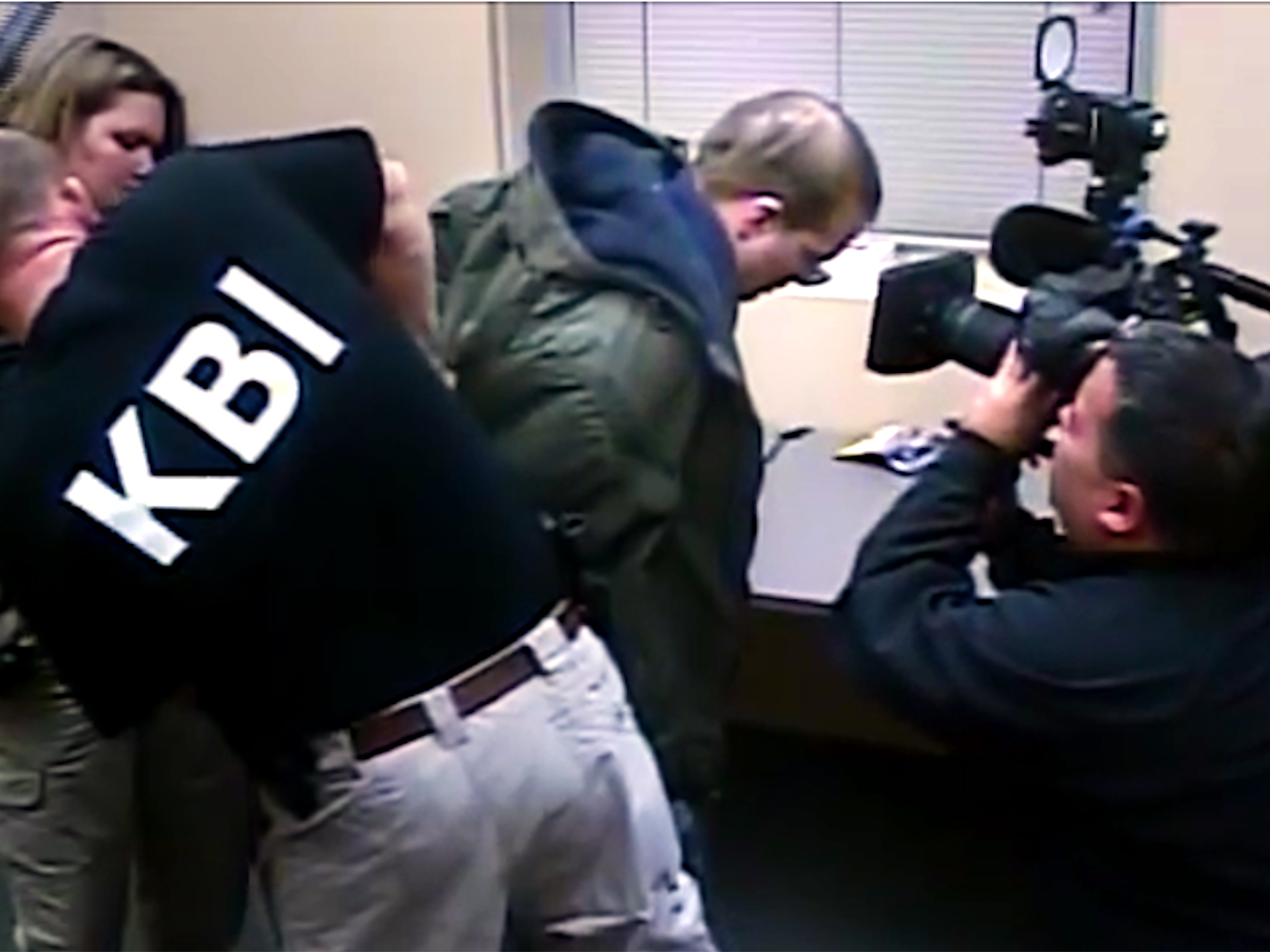 Man in green jacket being interviewed by journalists with cameras and microphones in office setting with venetian blinds.