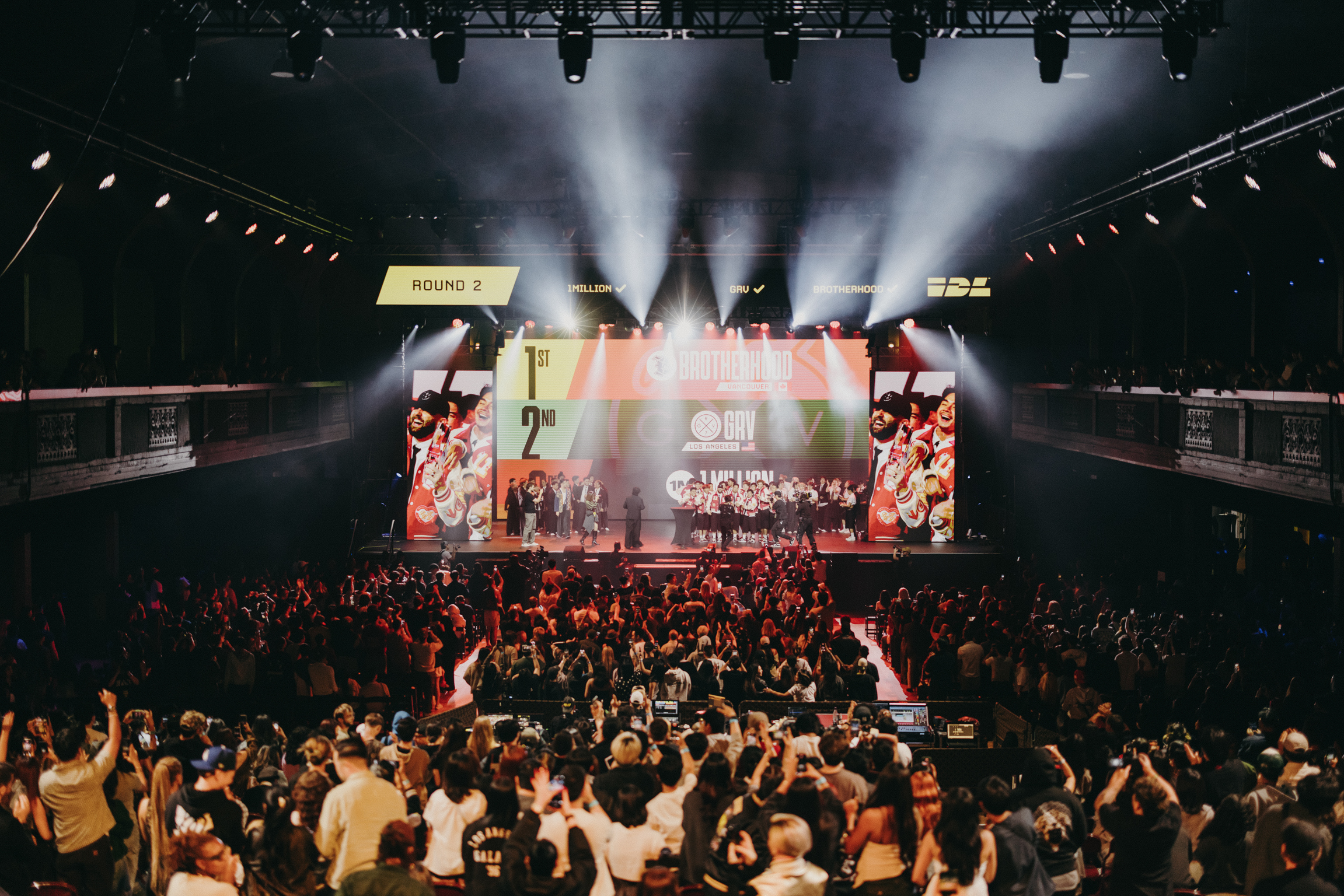 Large crowd in arena watching illuminated stage with bright spotlights, screens, and performers during live concert event.