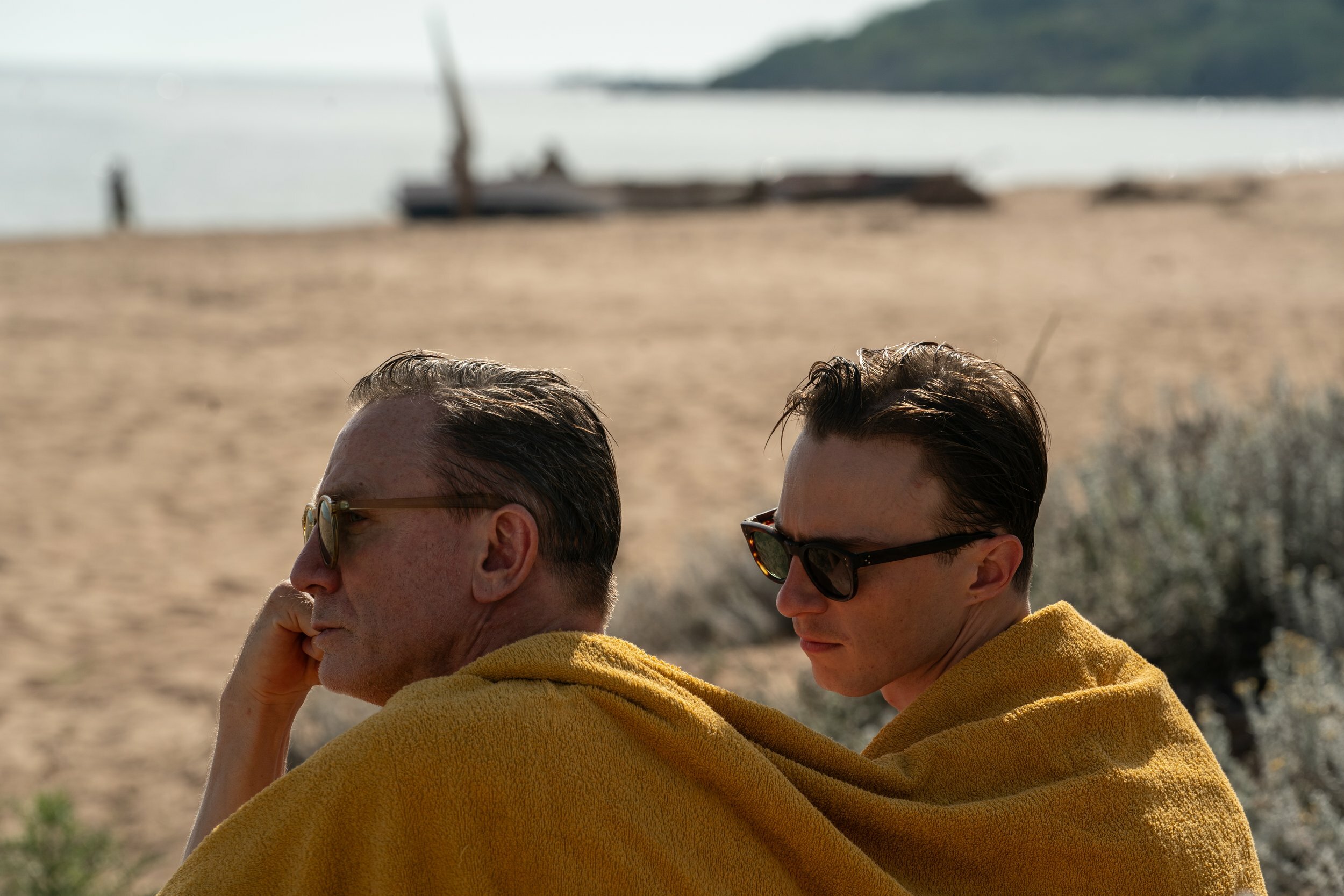 Two people wrapped in yellow blankets, facing each other on a sandy beach with a ship visible in the background.