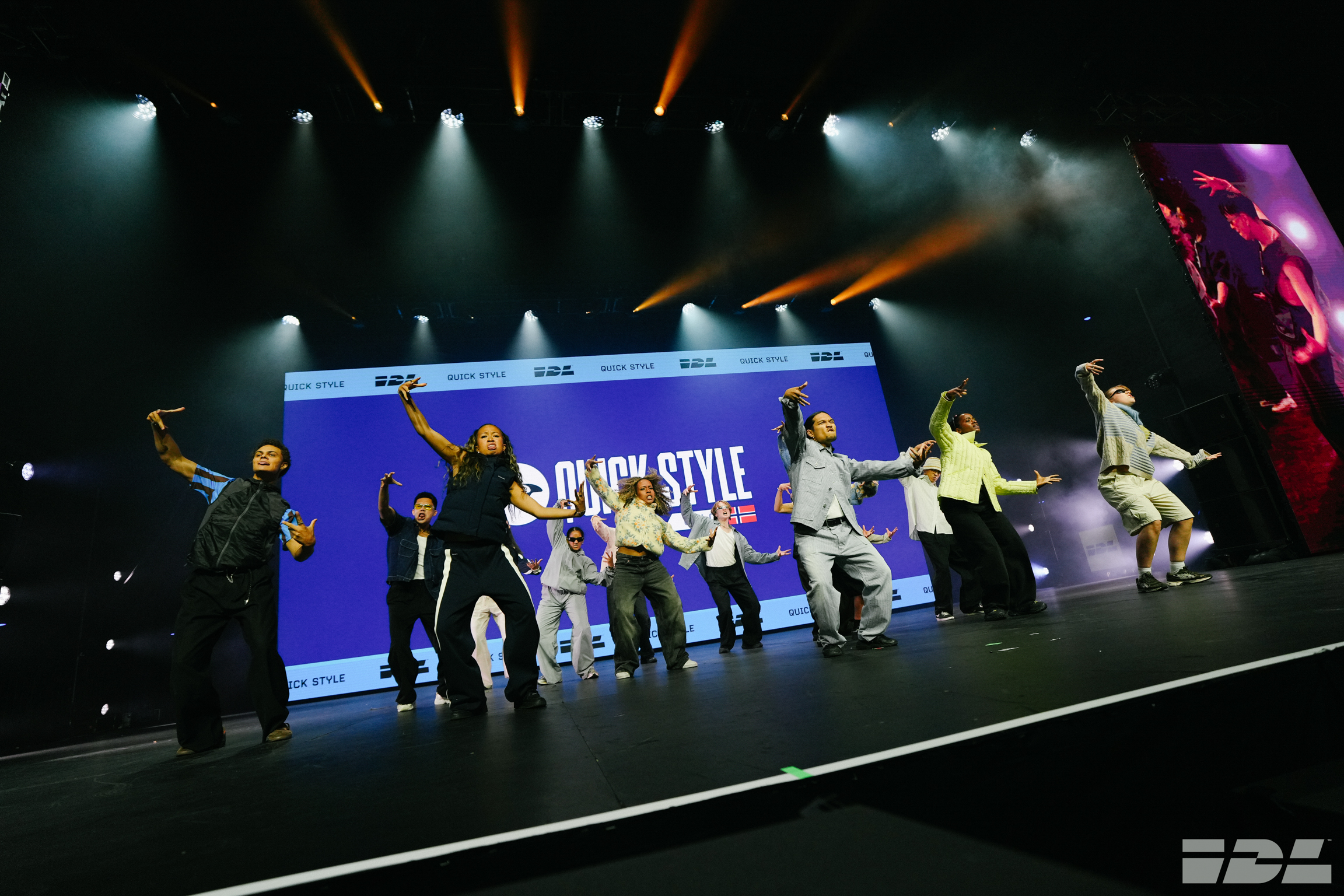 Group of performers on stage with arms raised, blue backdrop with "SIMPLY STYLE" text, colourful stage lighting from above.