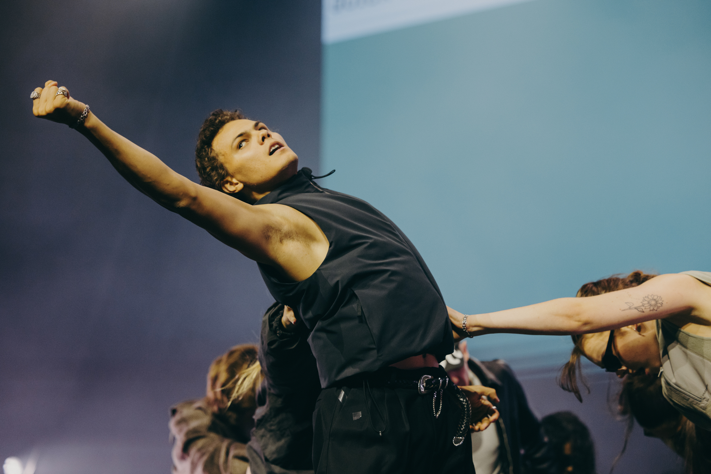 Female performer in black outfit with arm raised, other dancers visible below, blue-grey stage lighting background.