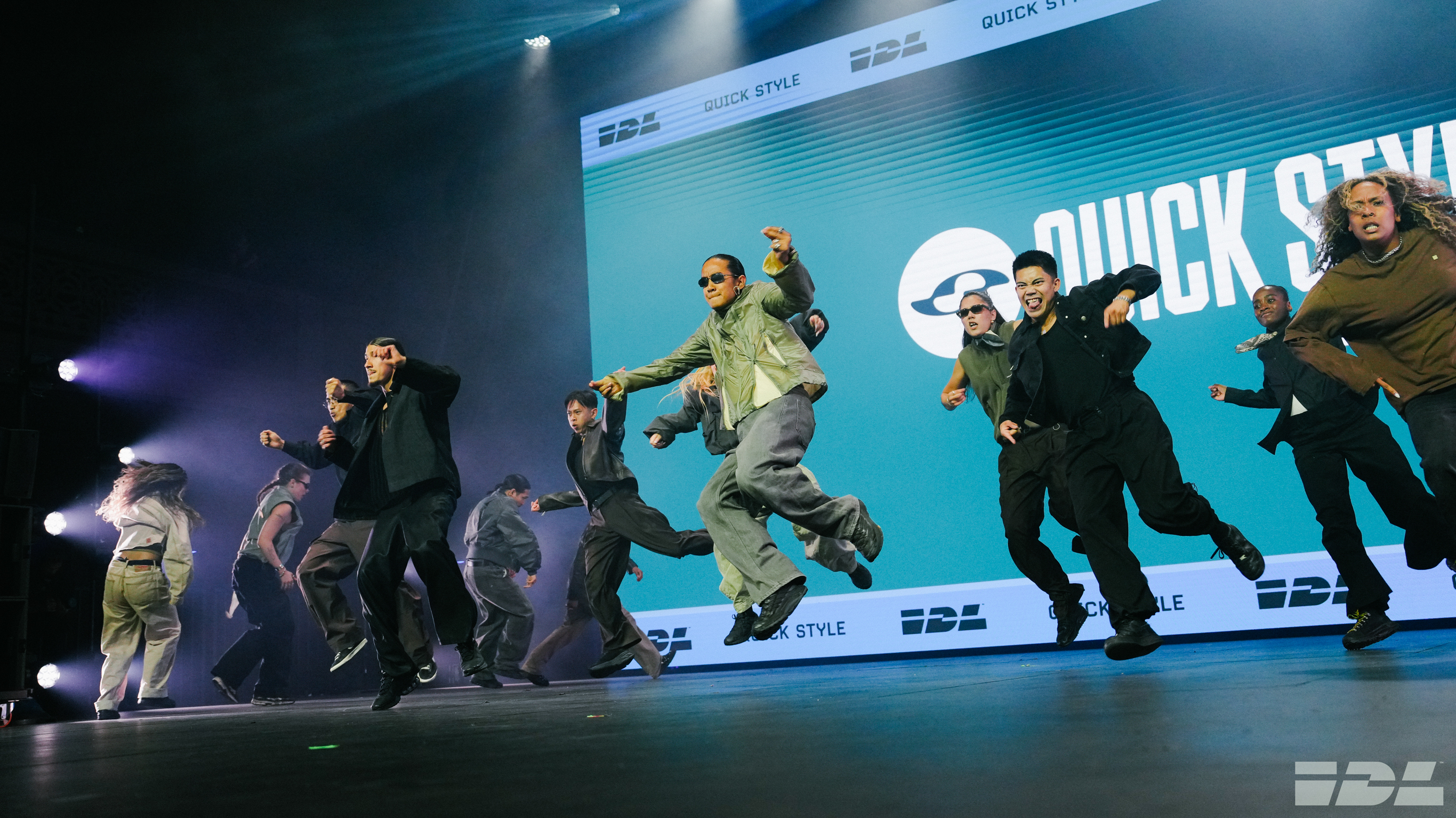 Dancers in military-style green and black clothing performing on stage with blue lighting and large screen displaying "QUICK ST" text.