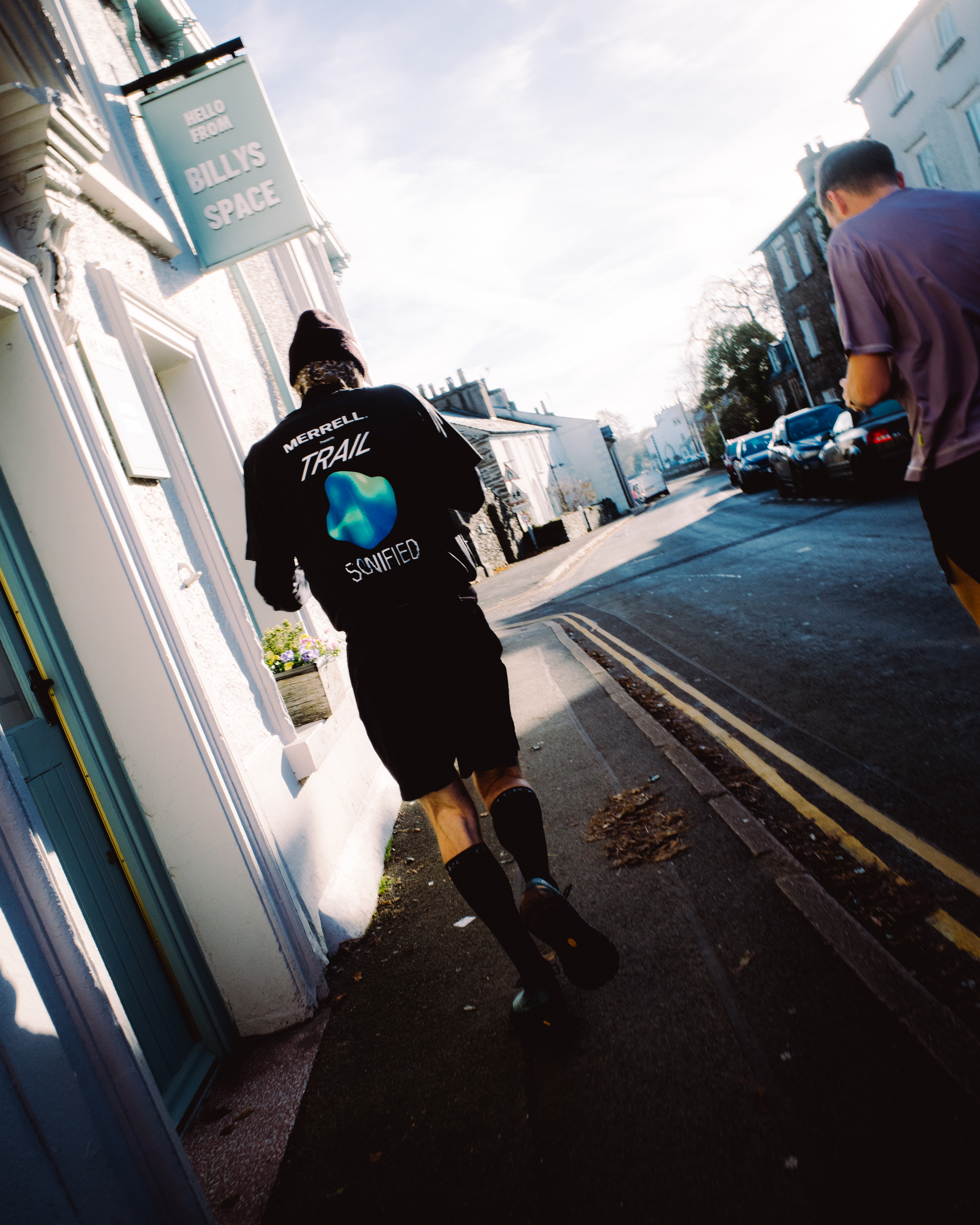 Person in black jacket walking on pavement past white shopfront with green sign, another person visible ahead on street.