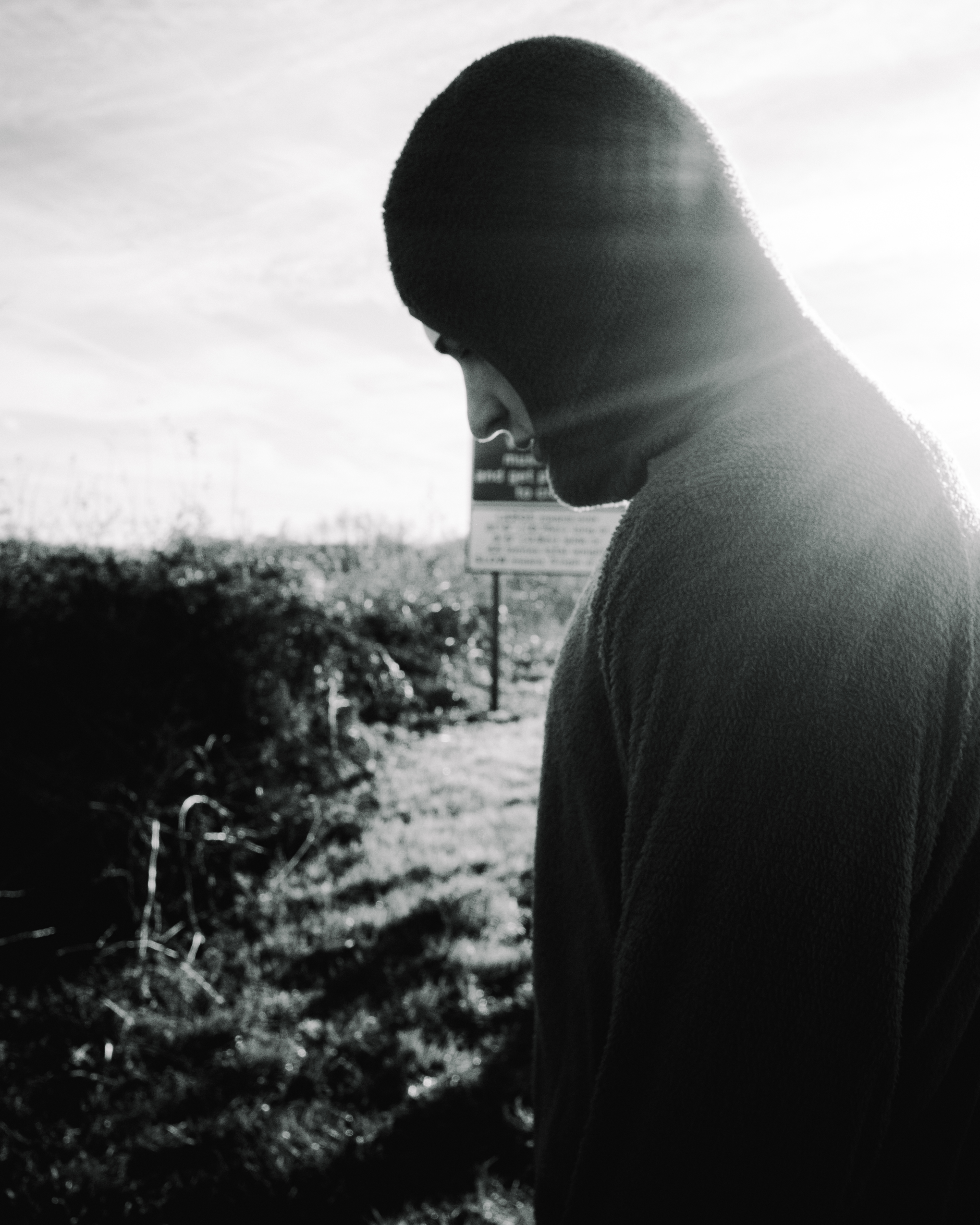 Man in profile looking down, standing outdoors near vegetation and wooden posts under cloudy sky. Black and white with green tint.