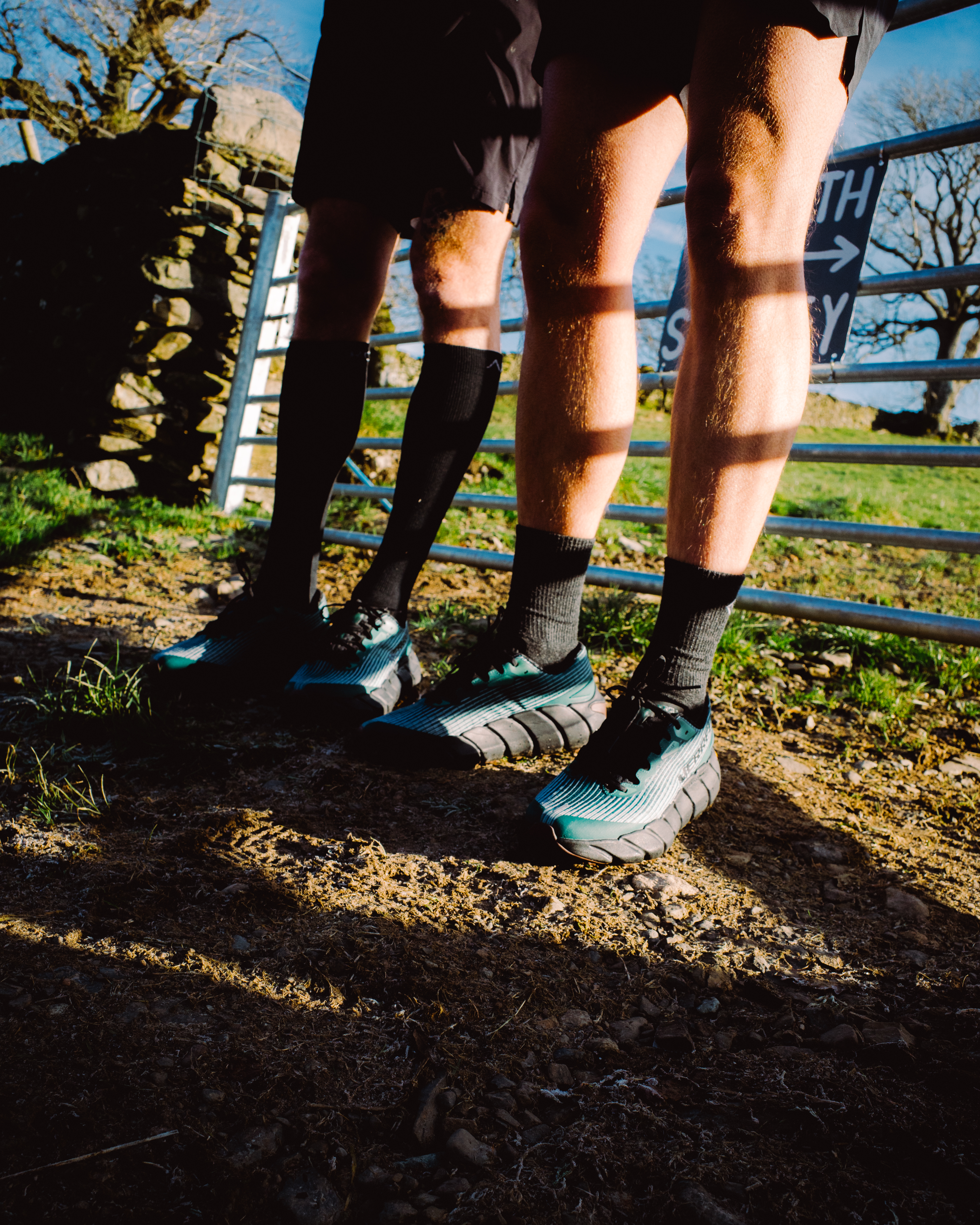 Two runners' legs wearing compression socks and trainers standing on muddy ground beside metal railings with trees in background.