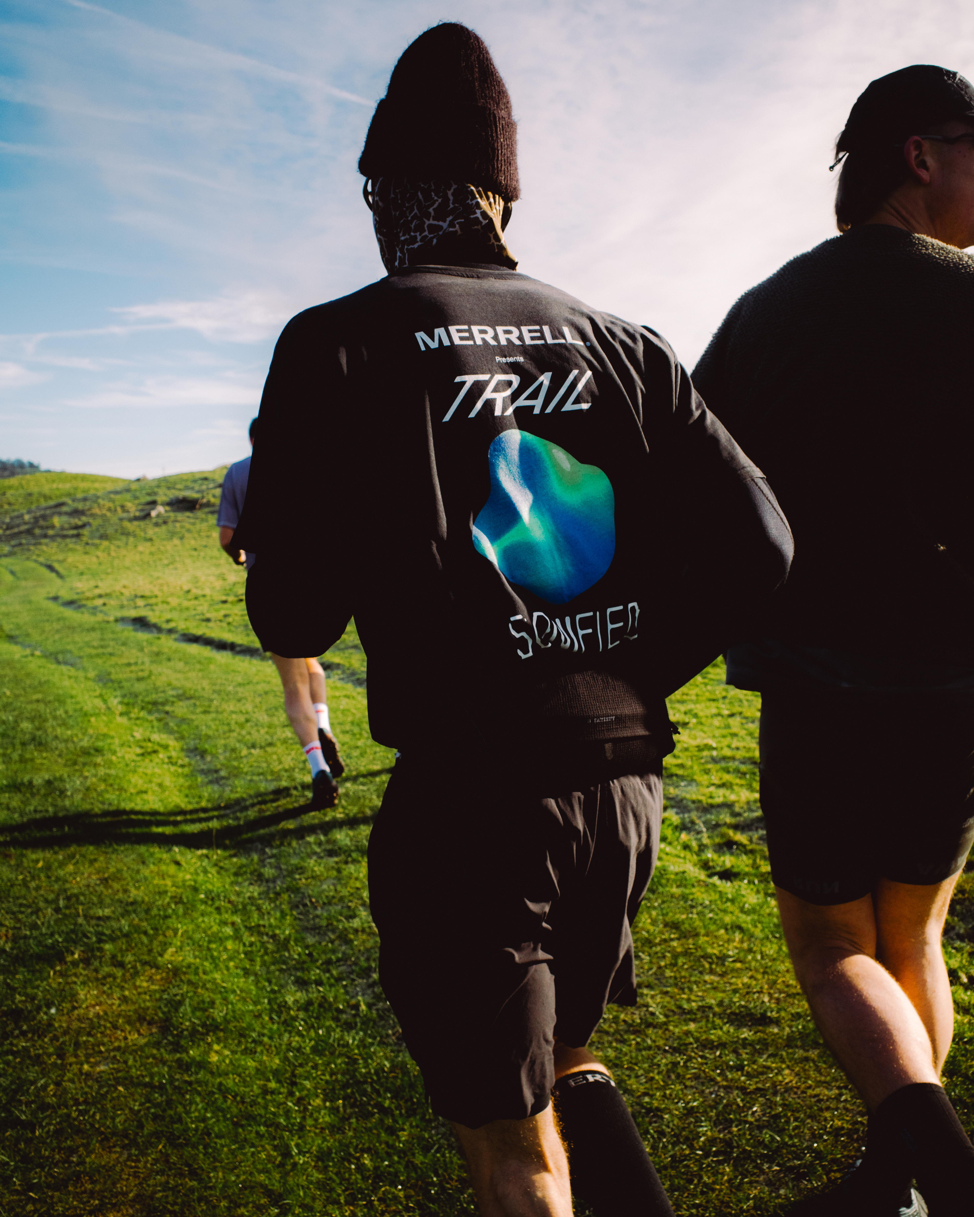 Two people running on green hillside, rear view. Person on left wears black jacket with "MERRELL TRAIL" text and blue-green logo.