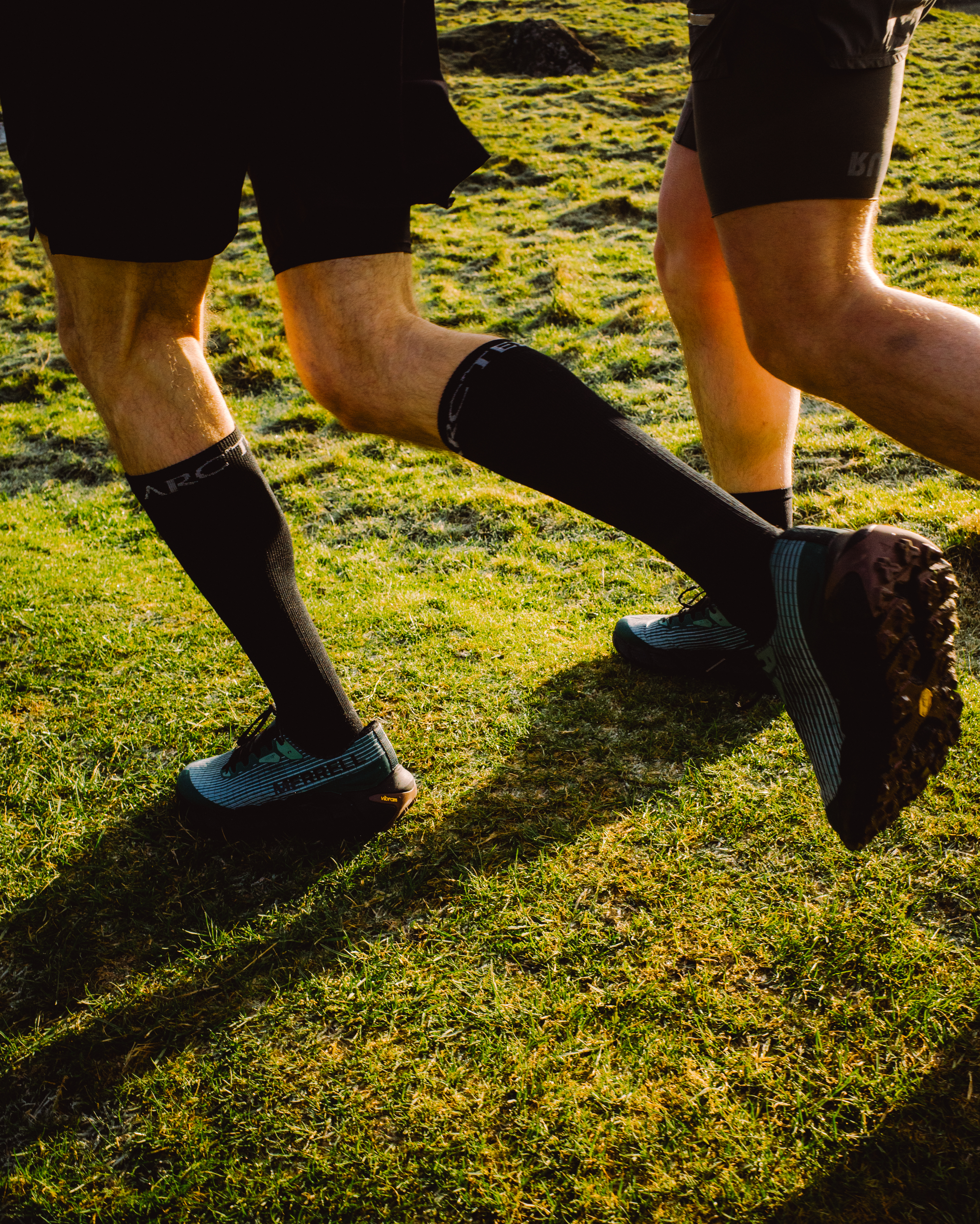 Two people's legs running on green grass, wearing black compression socks and dark trainers with blue accents in bright sunlight.